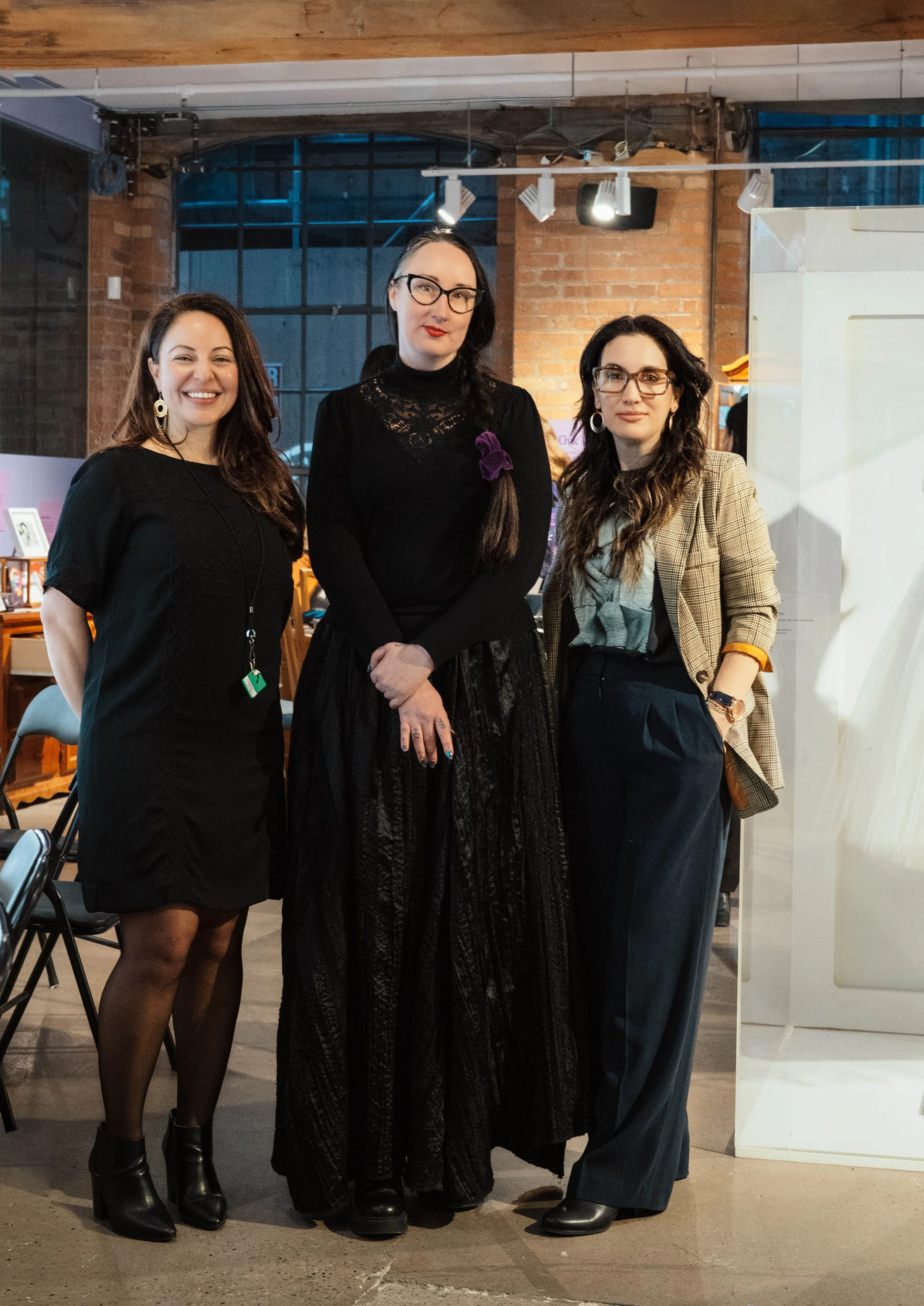 Three women standing together indoors at a social event, wearing fashionable attire, with a brick wall and artwork in the background.