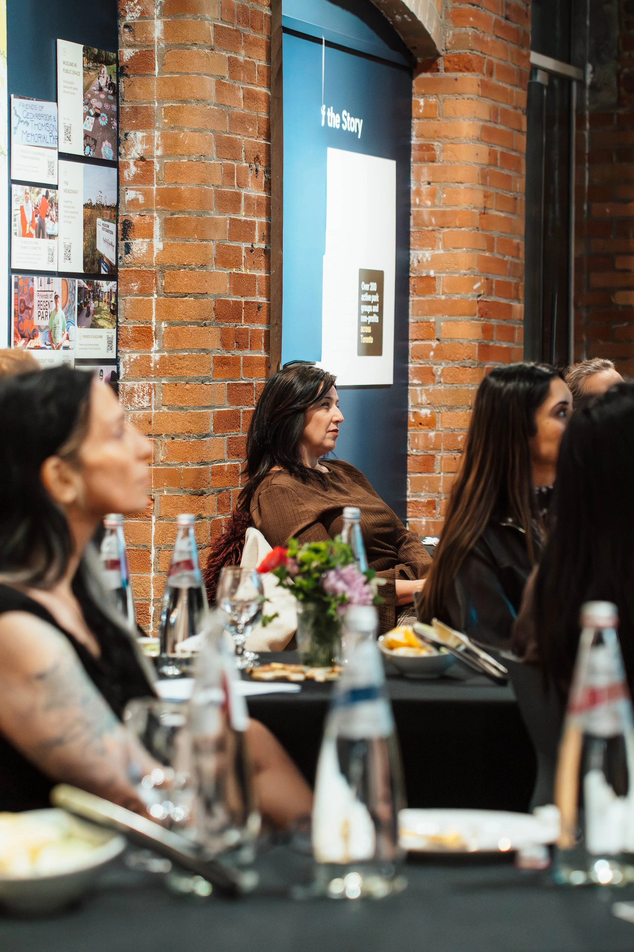 Women attending an event, sitting at a long table with plates, glass bottles, and a floral centerpiece inside a brick-walled room.