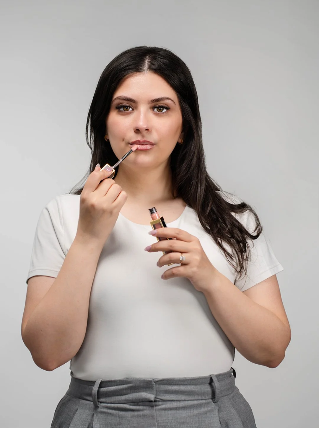 A woman with long dark hair applying lip gloss with a small brush while holding the lip gloss tube, wearing a white t-shirt and gray pants, against a plain gray background.