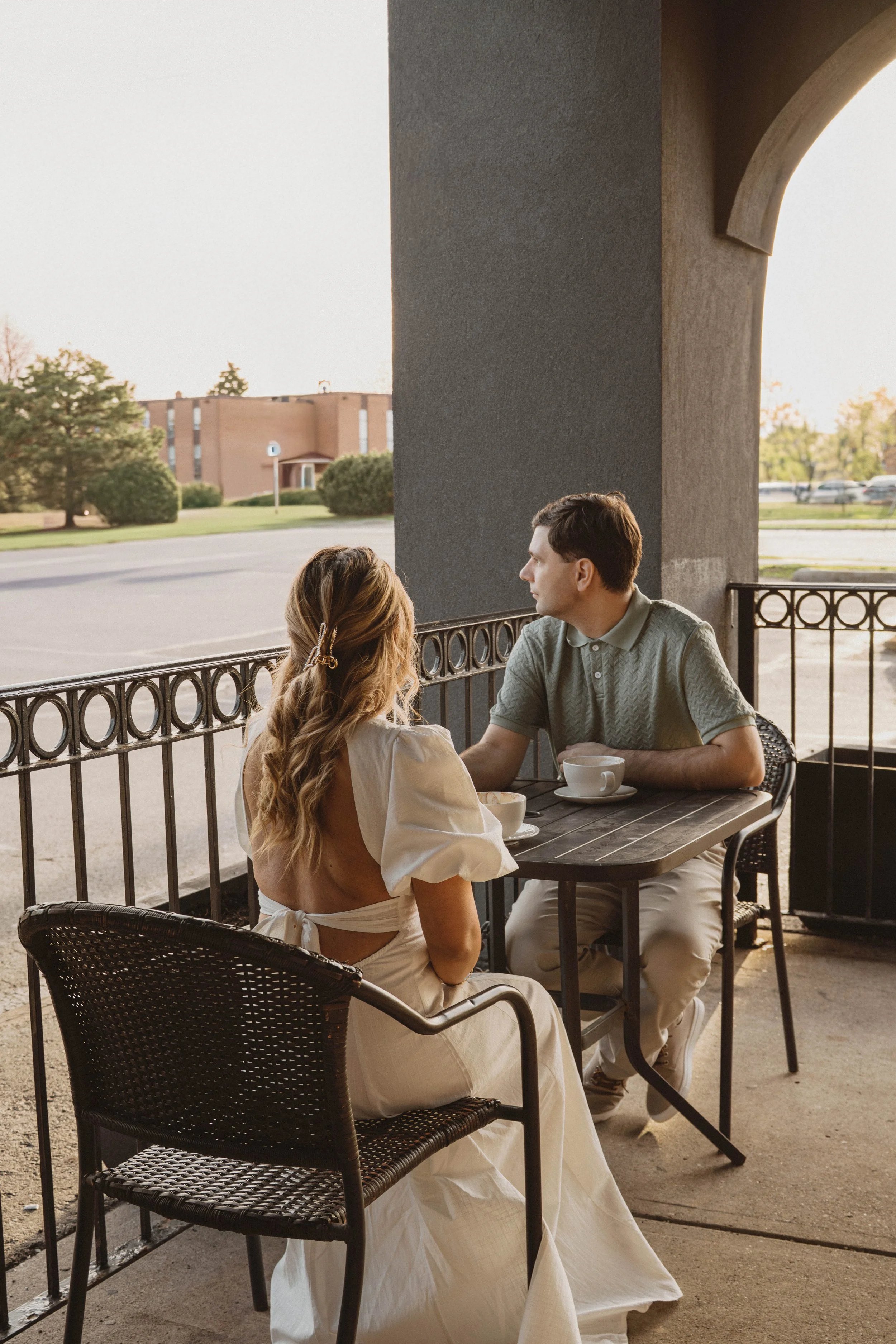 A woman with long, wavy hair and a hair clip, wearing a white dress, sitting at a table on a patio. A man with short brown hair, wearing a green polo shirt and light-colored pants, is sitting across from her. They are having coffee, with cups on the 