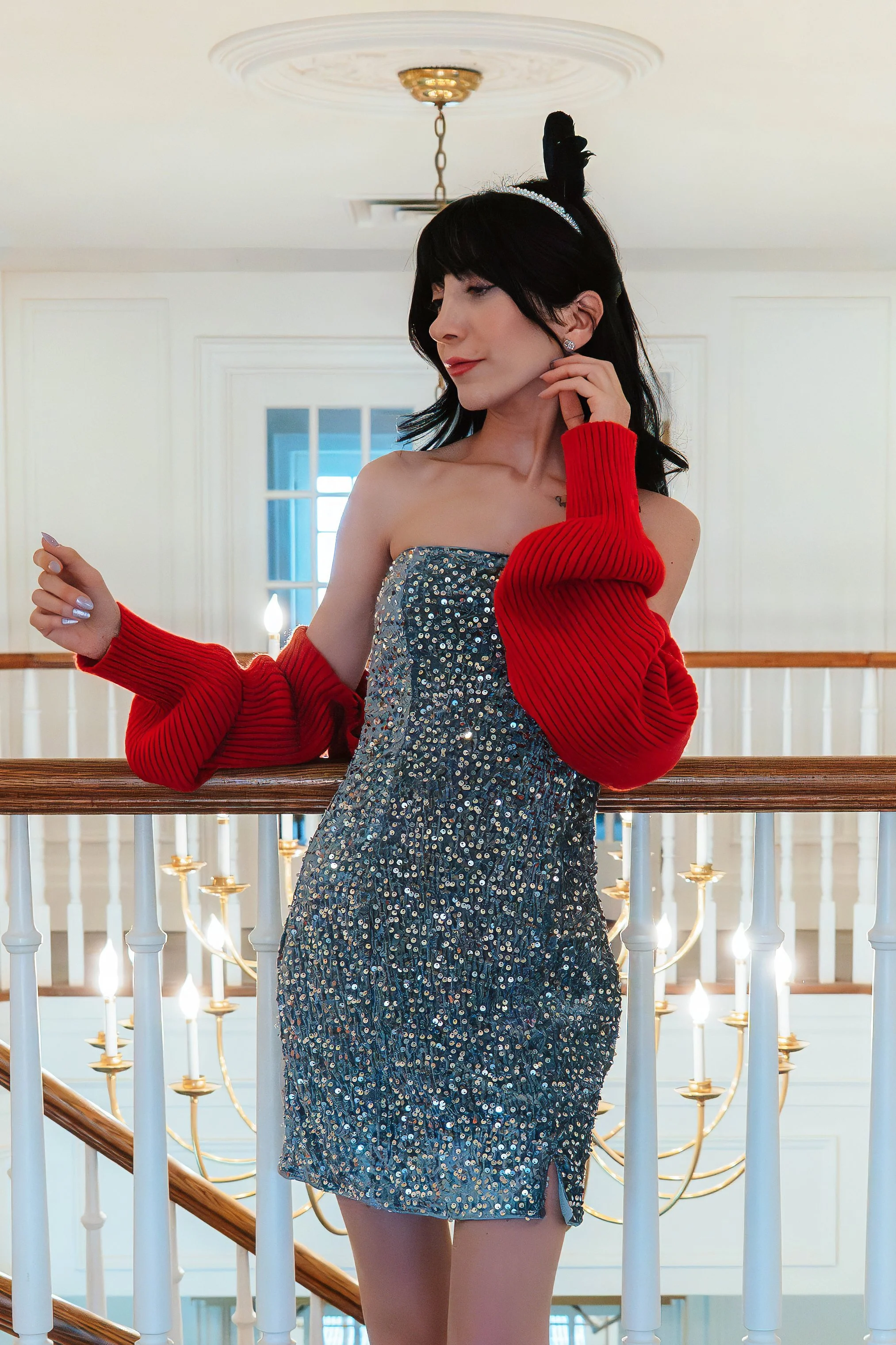 Woman standing on a staircase with a railing, wearing a glittery dress and red sweater sleeves, with a black headpiece and pearl earrings, inside a well-lit interior with chandeliers and a blue door or window in the background.