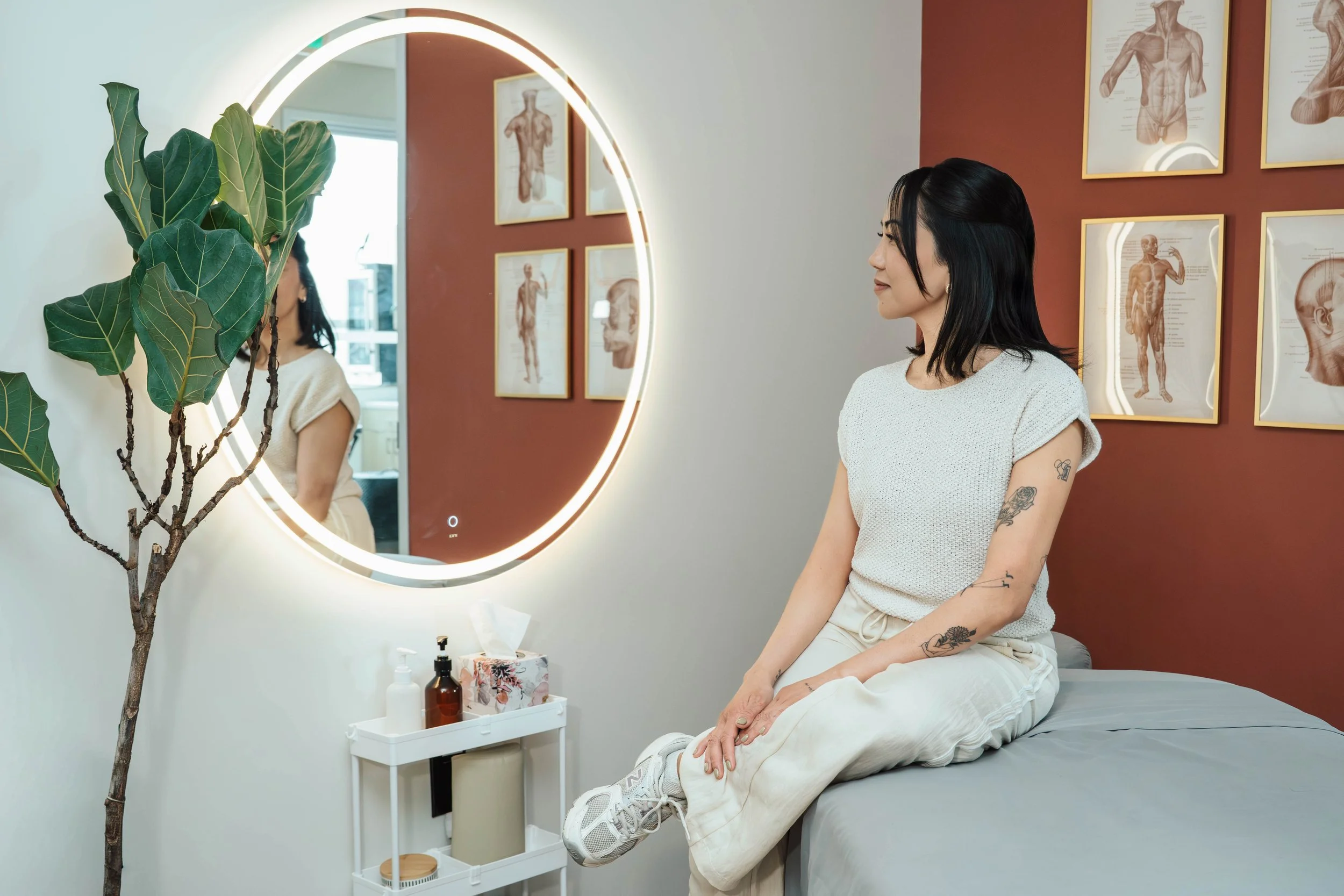 A woman with black hair, tattoos on her arm, dressed in a white top and beige pants, sitting on an examination table in a room with medical charts on the wall, looking at herself in a circular mirror with LED lighting, a potted plant in the foregroun