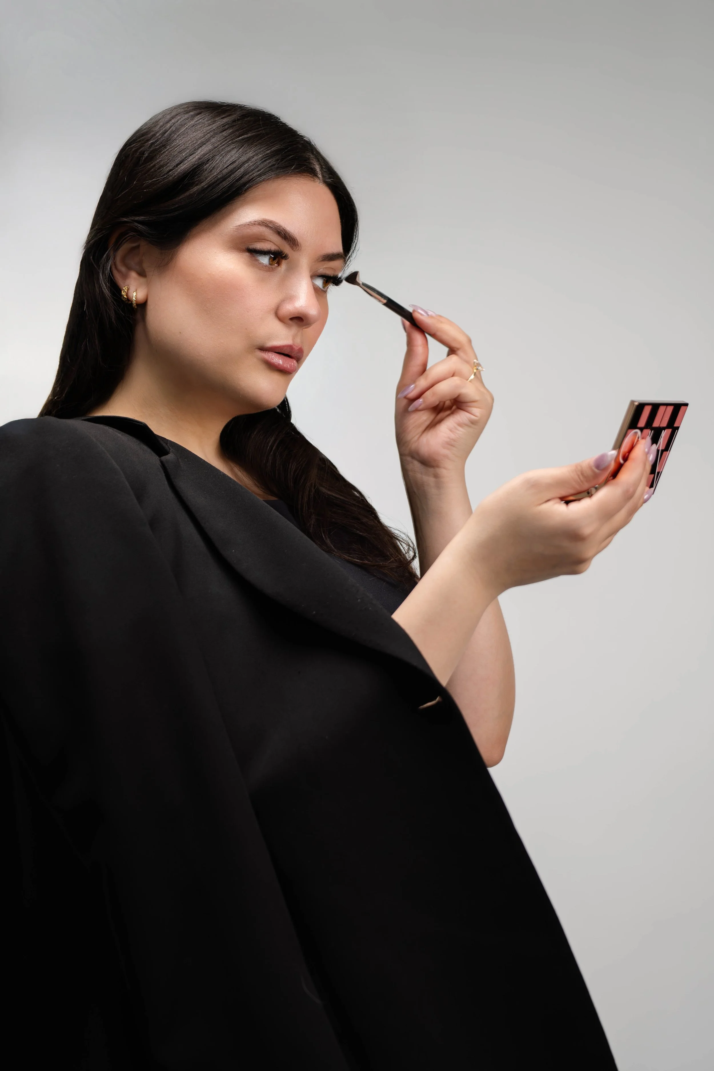 A woman with long dark hair and earrings applying makeup with a brush while holding a mirror in her other hand.
