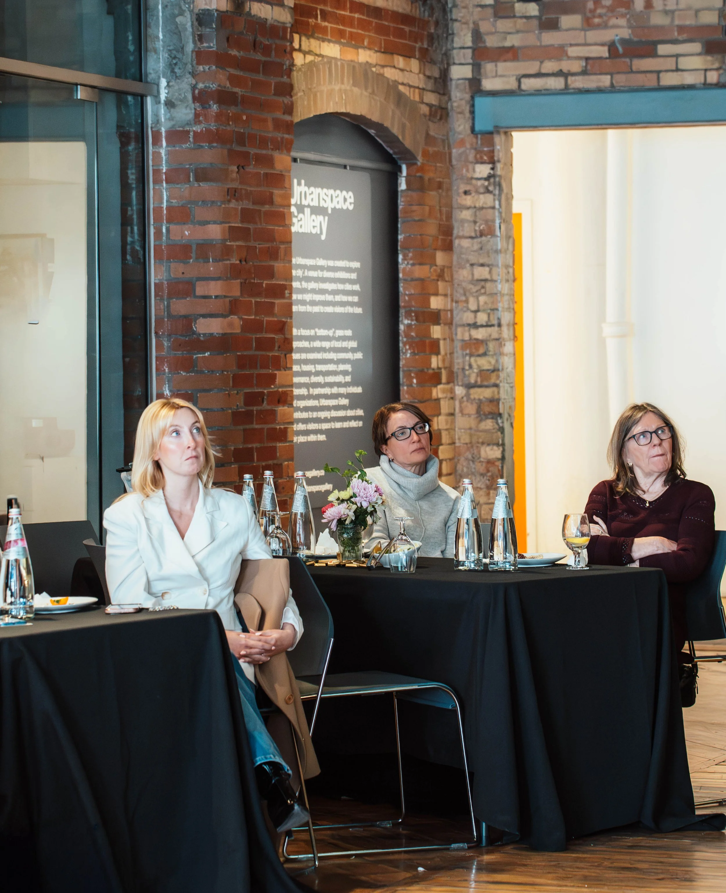 Three women sitting at a table with bottled water and glasses, listening attentively during a presentation or discussion in an art gallery with exposed brick walls.