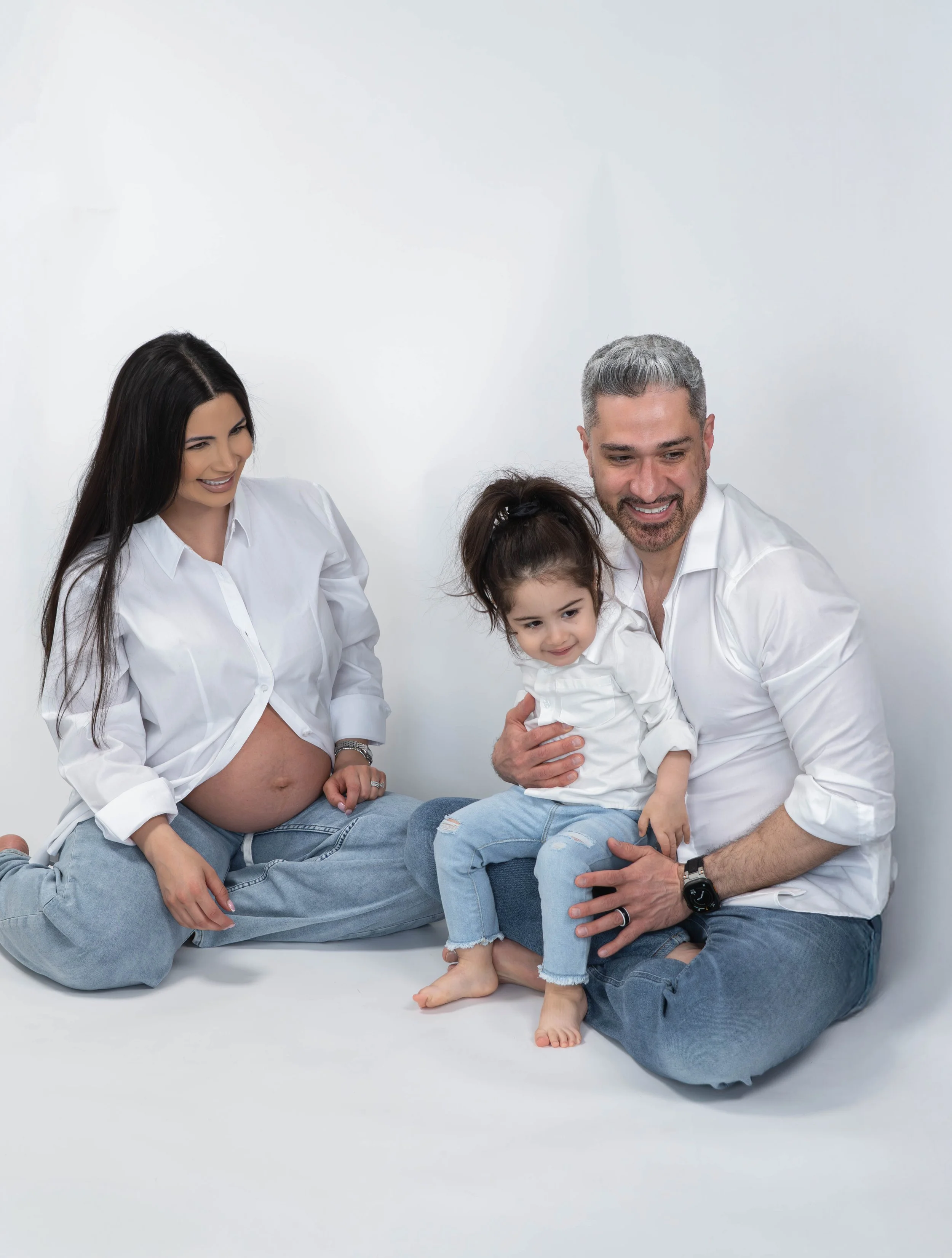 A pregnant woman, a man, a young girl, and a woman with long dark hair are sitting together on a white background, smiling and looking at each other, dressed in white shirts and casual jeans.