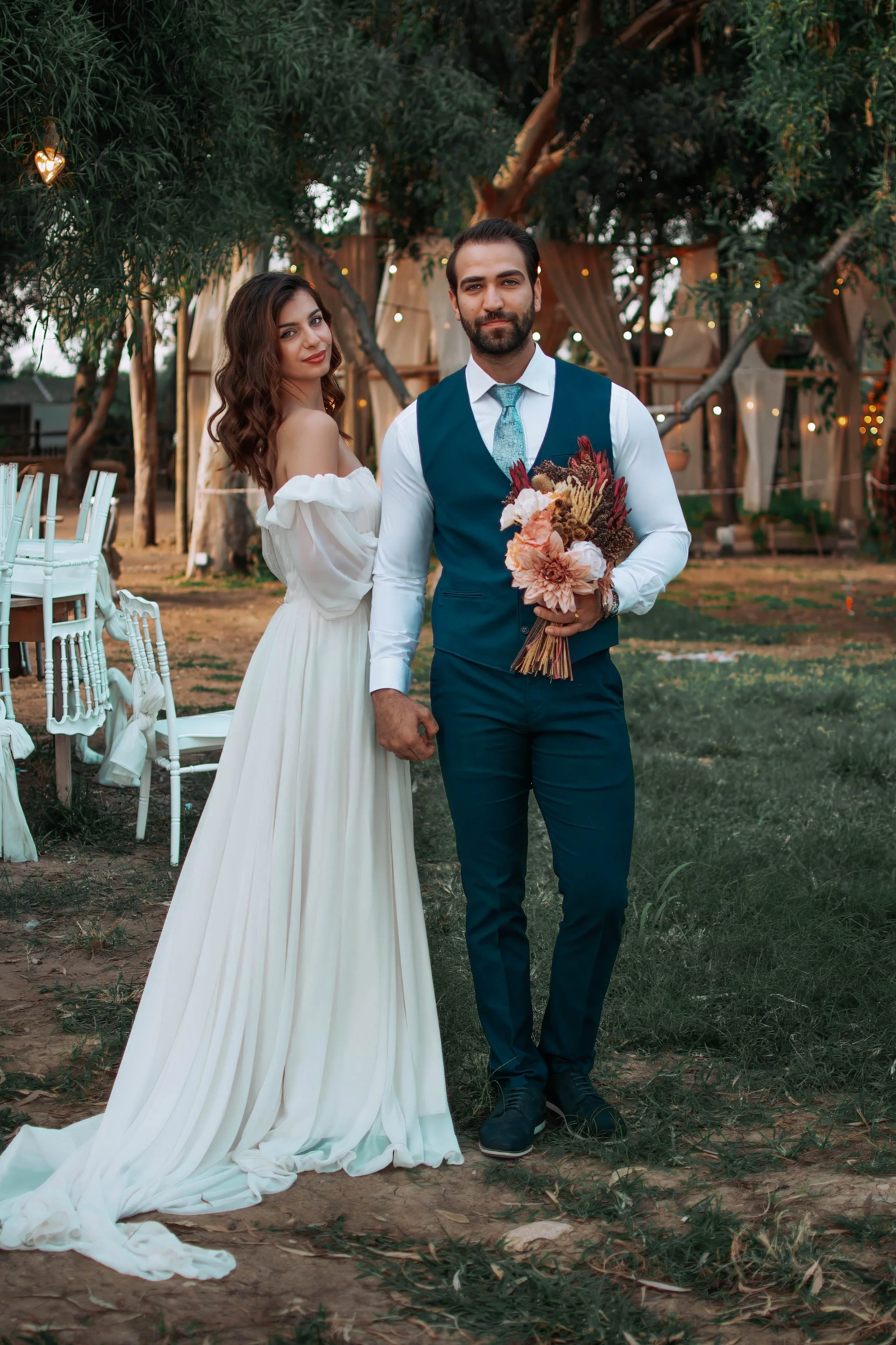 A bride and groom pose for a wedding photo outdoors during daytime. The bride wears a vintage off-shoulder white gown with a long train, and the groom is dressed in a blue vest, white shirt, and tie, holding a bouquet of flowers. They are standing on