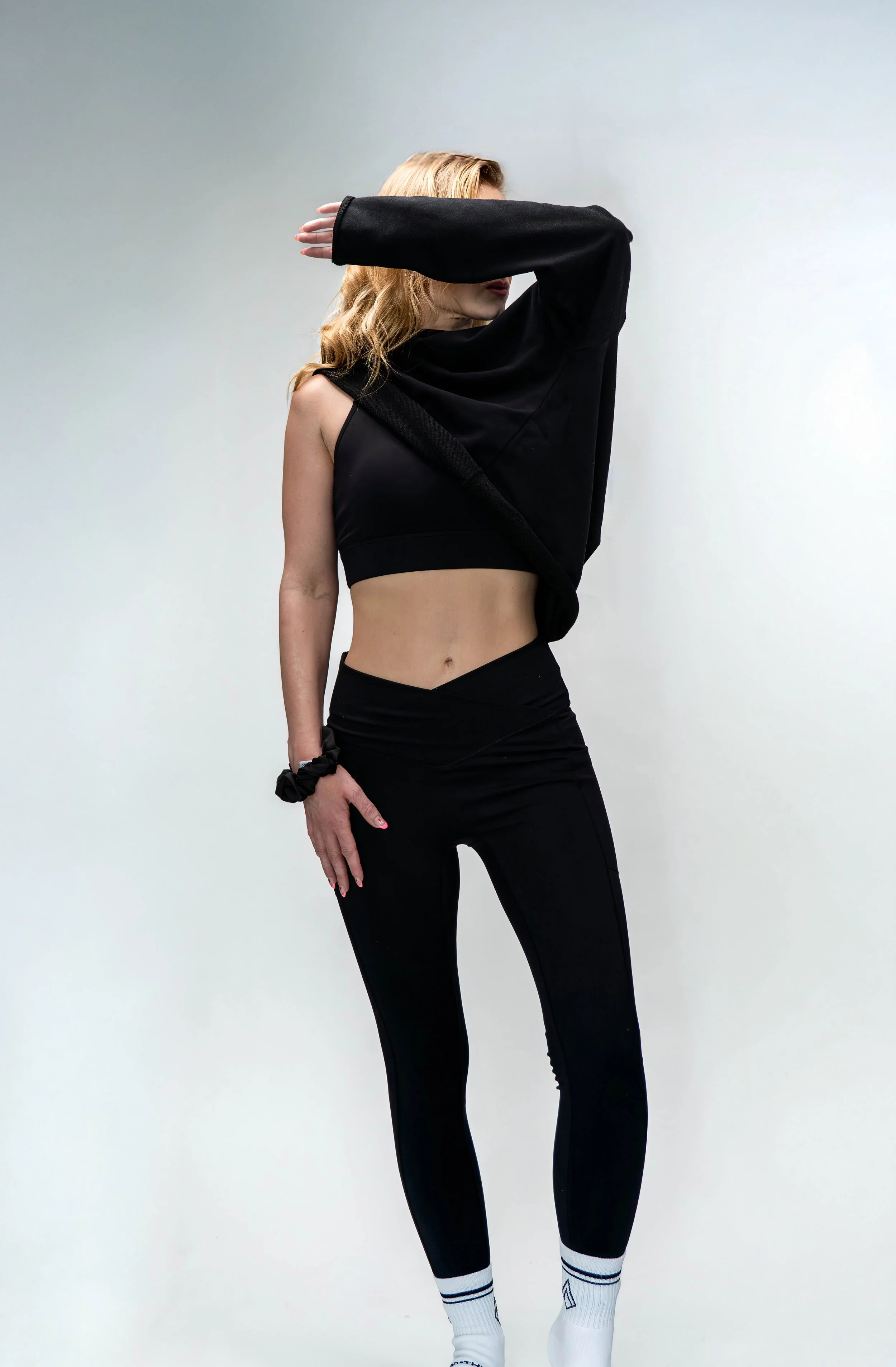 A woman with wavy blonde hair wearing a black athletic crop top, black leggings, and white sports socks posing against a plain light-colored background.