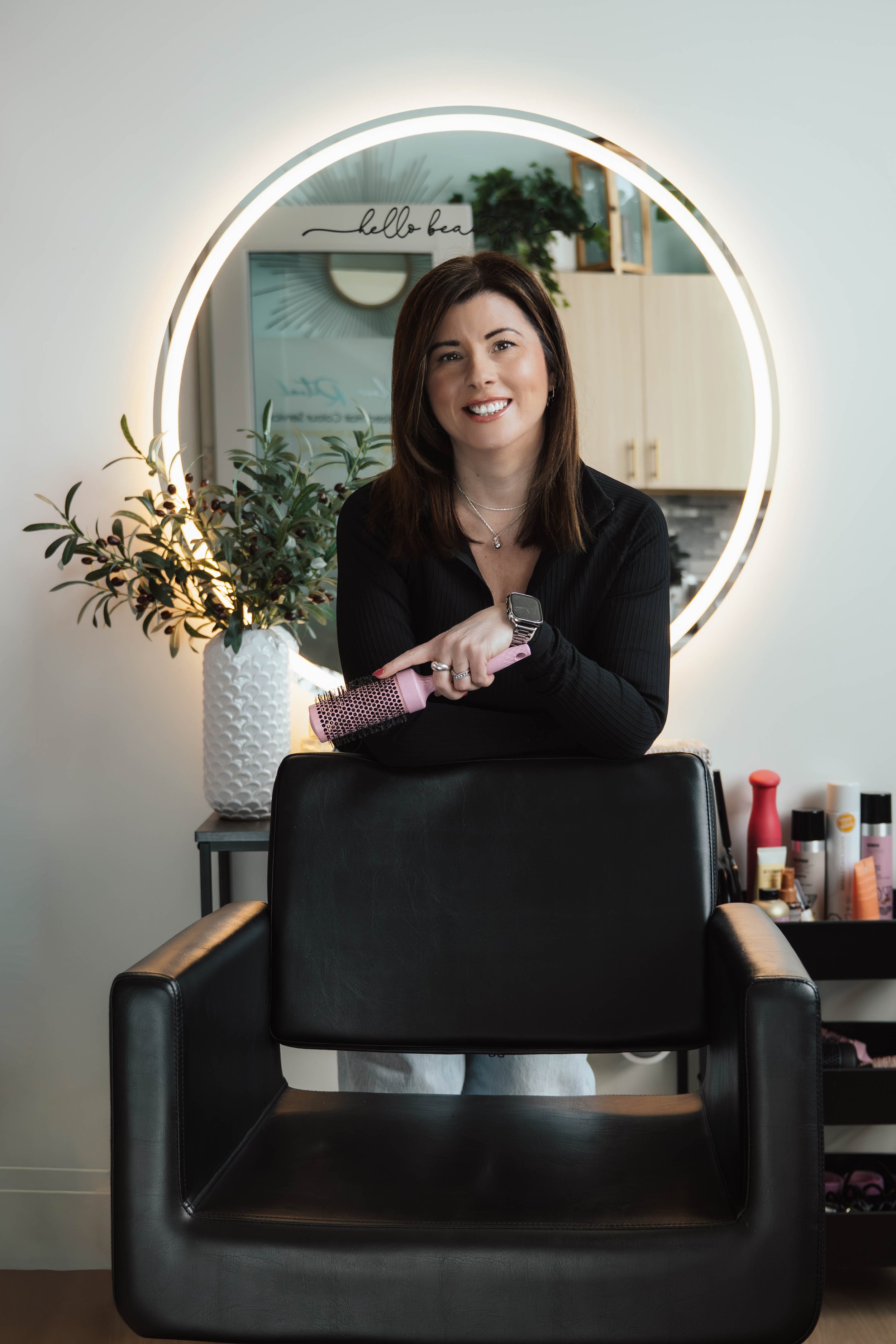 Woman standing behind a salon chair, holding a pink round hairbrush. She is smiling at the camera, with a round mirror and decorative plant behind her. Salon supplies are visible on a cart to the right.