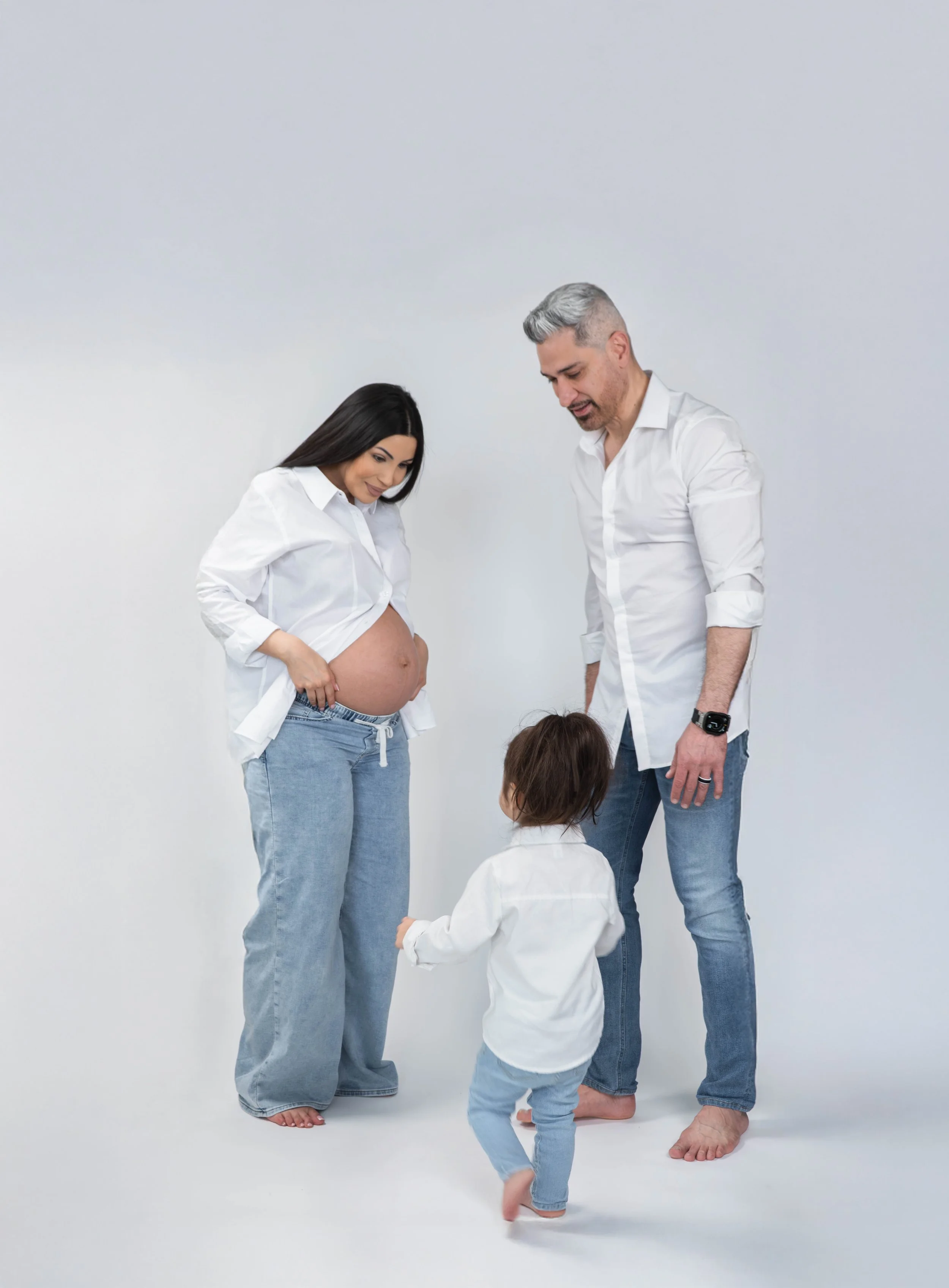 A pregnant woman in a white shirt and jeans standing next to a man in a white shirt and jeans, both looking at a young girl in a white shirt and jeans who is holding the pregnant woman's belly, in front of a plain white background.