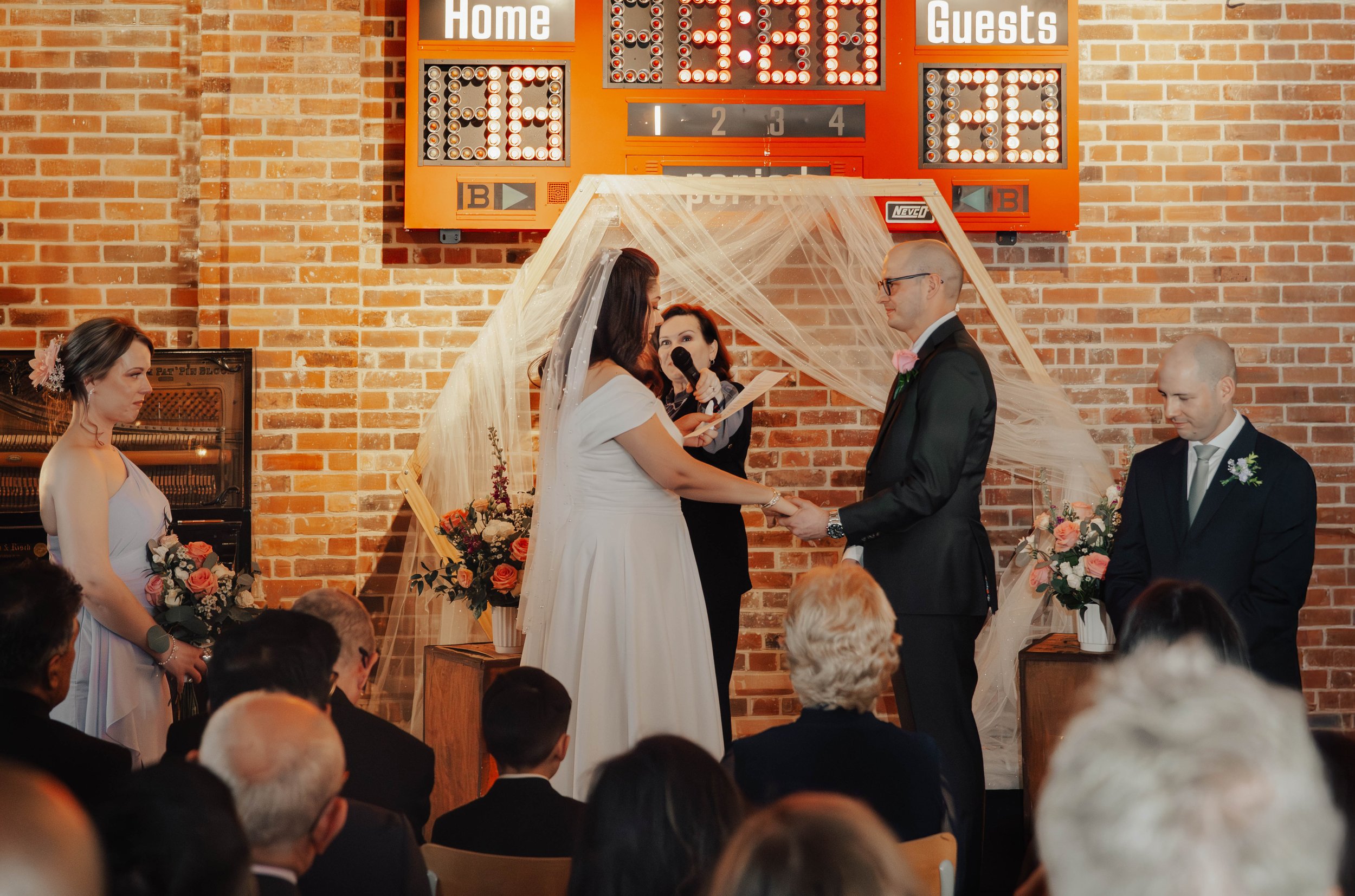 A wedding ceremony taking place indoors in front of a brick wall with a scoreboard. The bride and groom are holding hands, facing each other under a sheer fabric canopy. The officiant is reading from a paper, and the bridesmaid and groomsman stand ne