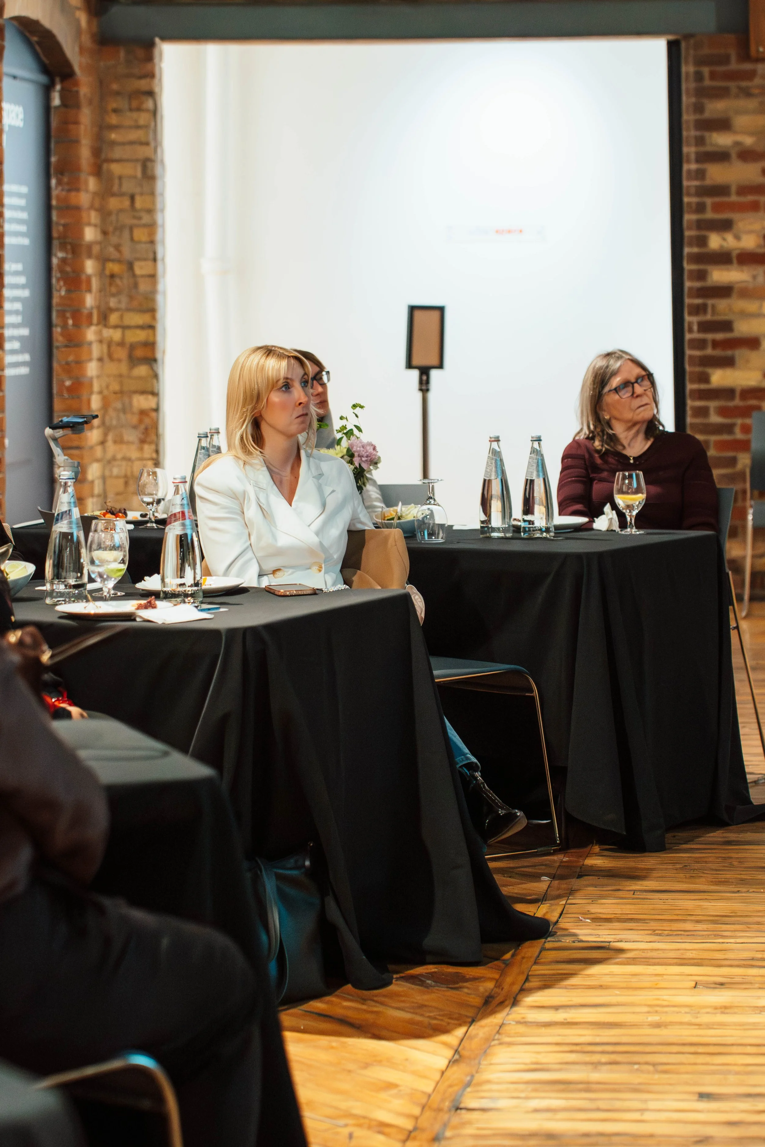Women attending a conference or seminar, sitting at black-covered tables with water bottles, glasses, and plates, in a room with brick walls and wooden flooring.