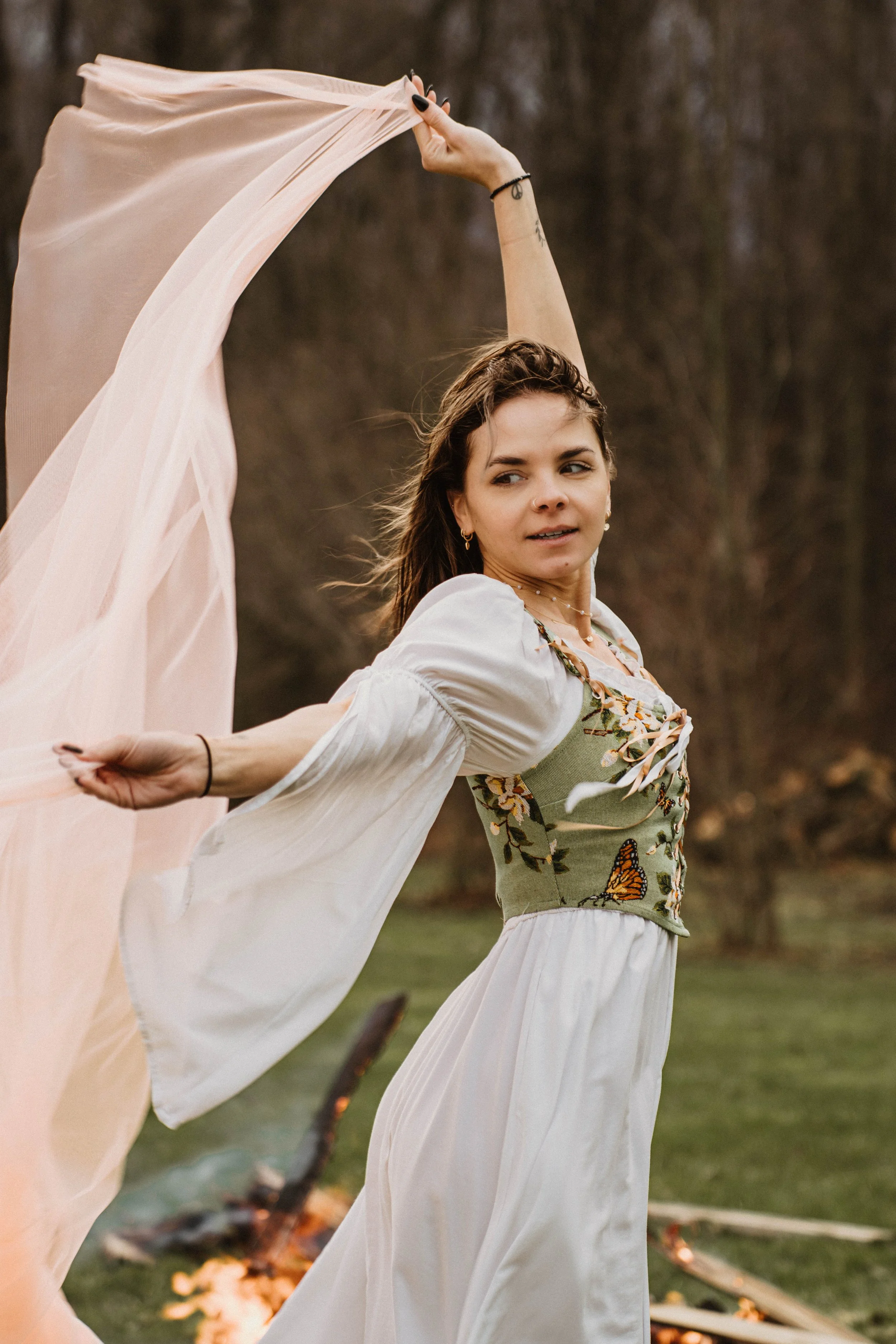 A young woman wearing a bohemian style dress with embroidered butterflies and flowers, holding a flowing pink fabric in an outdoor setting with a fire on the ground and trees in the background.