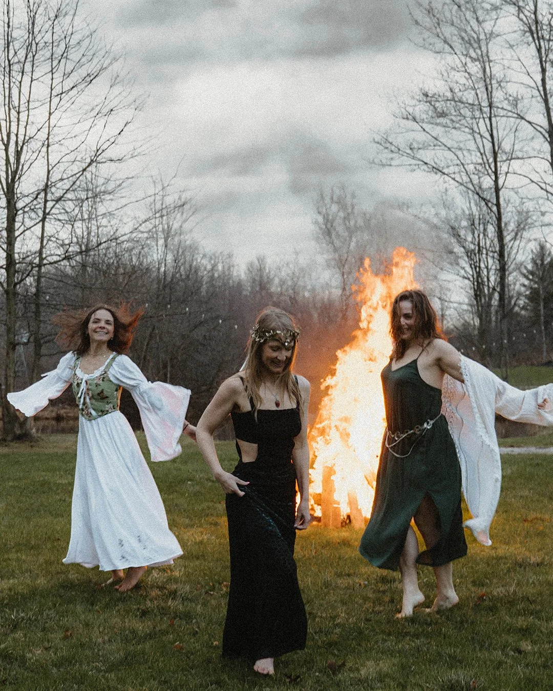 Three women dancing outdoors near a bonfire on a cloudy day, wearing bohemian-style dresses.