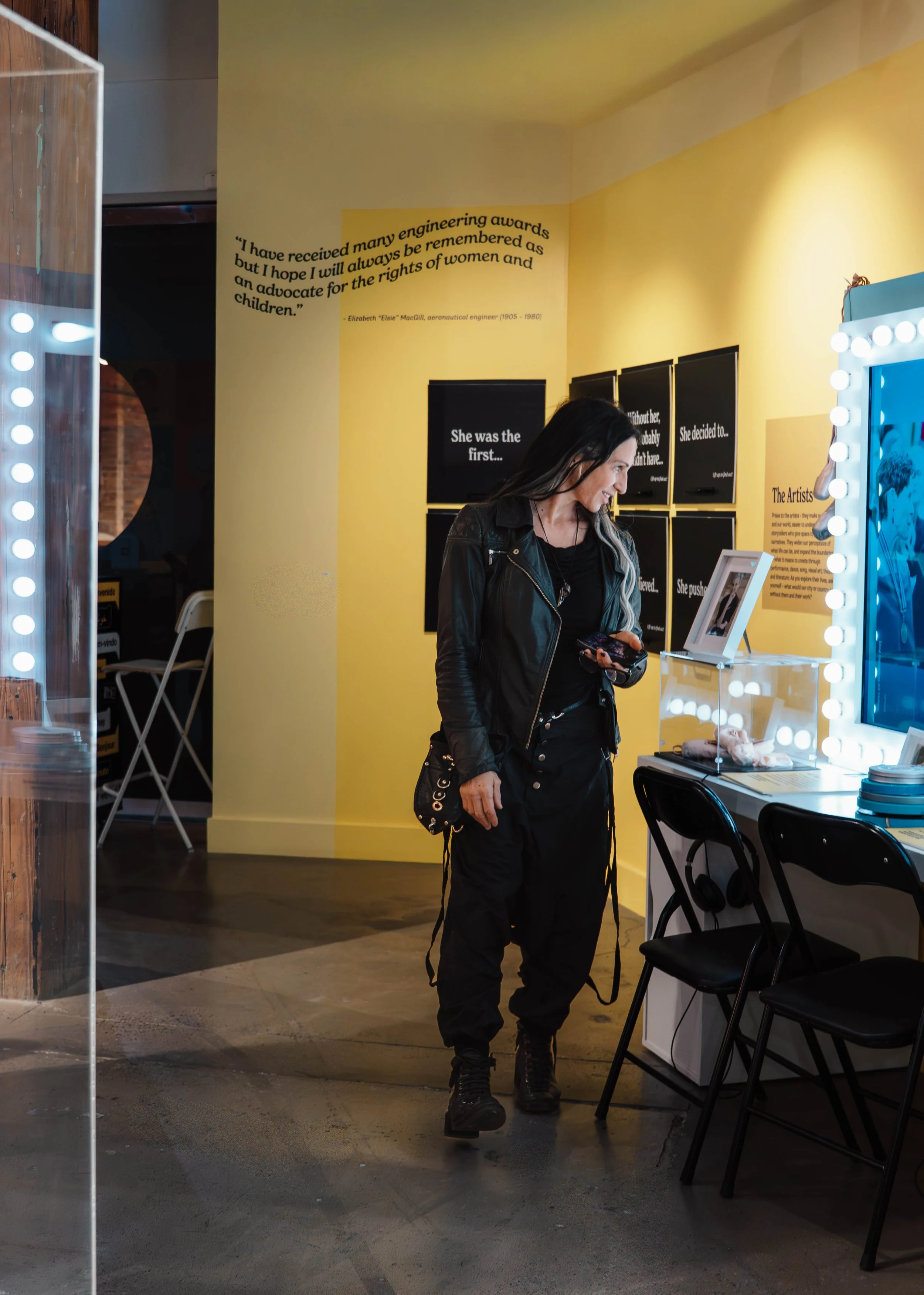 A woman dressed in black standing in an art gallery or exhibition space, looking at her phone, with yellow walls decorated with quotes and artwork behind her.
