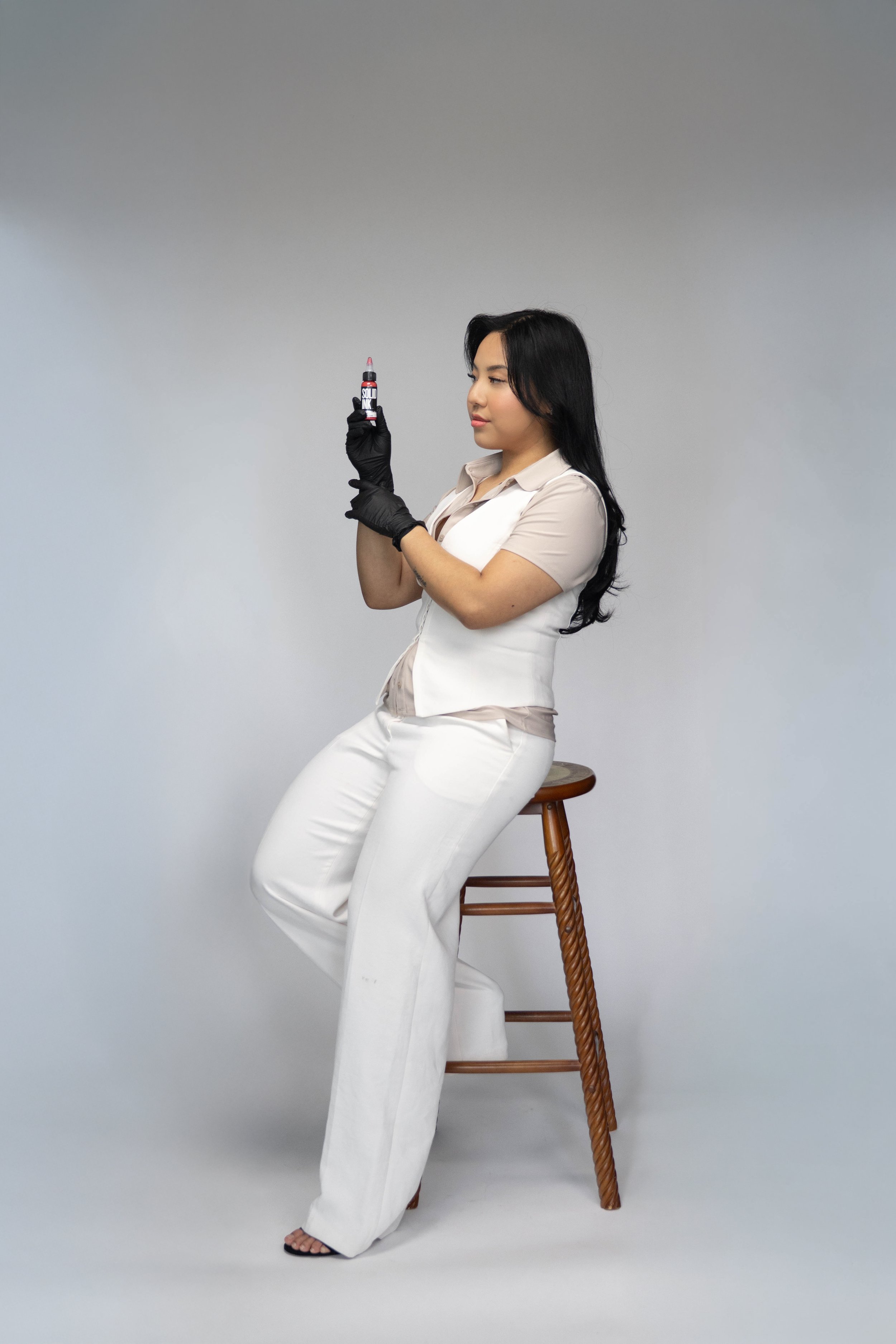 A woman with long black hair wearing a white outfit and black gloves, sitting on a wooden stool, holding a tube of lip gloss or lipstick and looking at it.