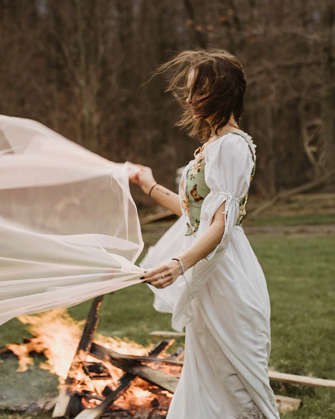 A woman in a dress with a butterfly pattern stands outdoors near a campfire, holding a sheer cloth.