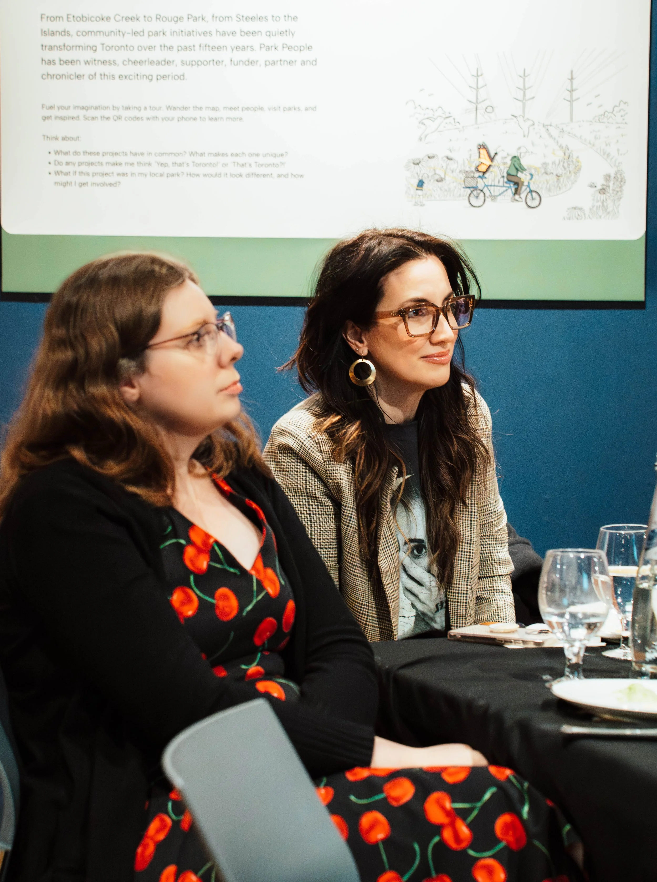 Two women sitting at a table listening attentively, with a presentation slide on the wall behind them.