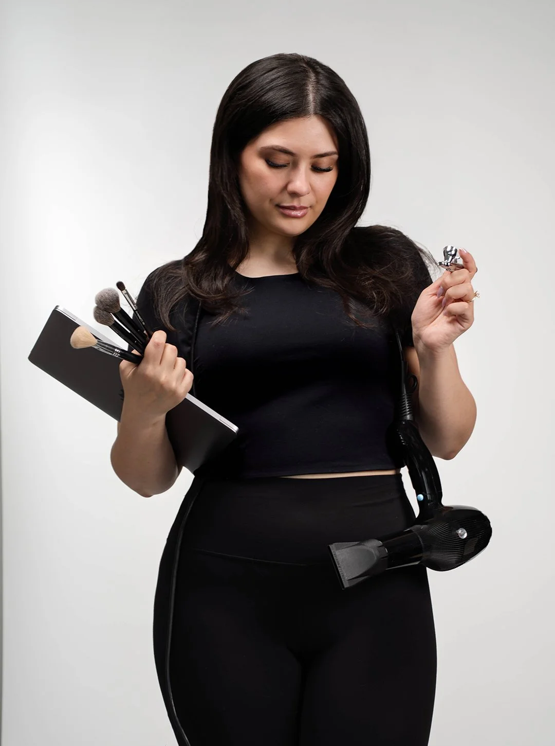 A woman holding makeup brushes and a notebook in one hand, and a small makeup tool in the other, with a vacuum cleaner hanging around her waist.