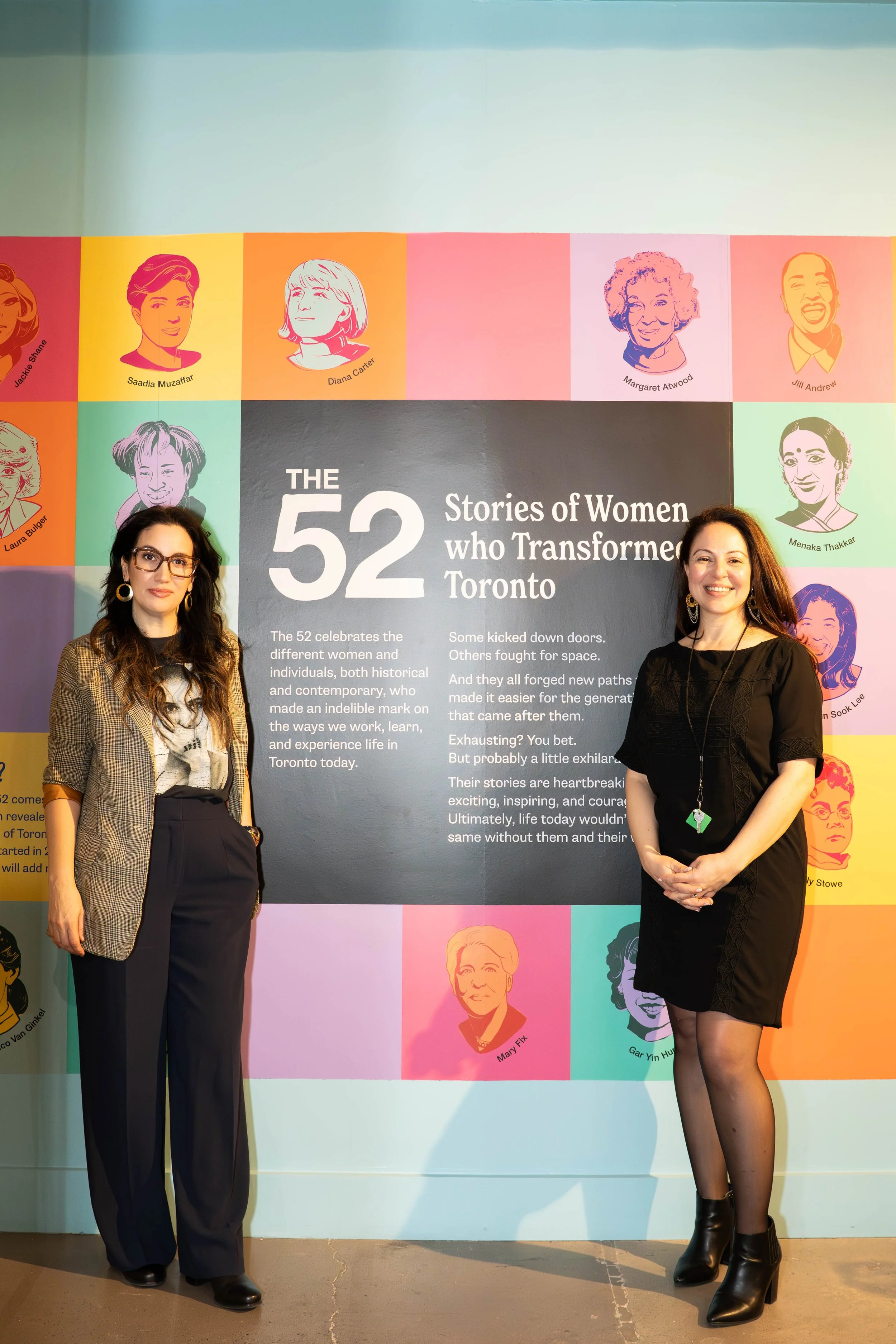 Two women standing in front of a colorful mural titled 'The 52 Stories of Women who Transformed Toronto'. The mural features portraits and names of various women, with a large black central section containing white text describing the project.