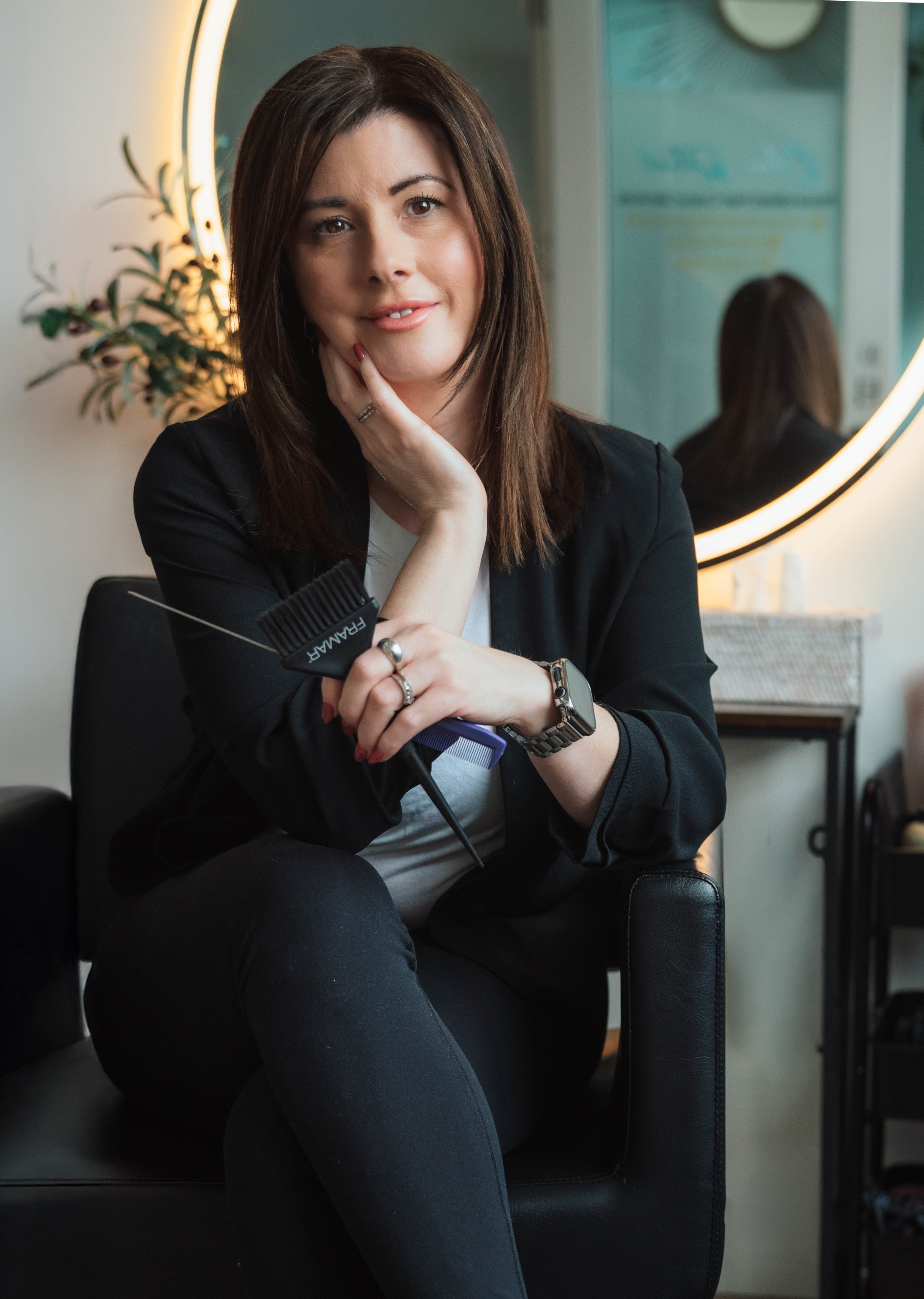 A woman sitting in a chair, holding a hairbrush and comb, with her other hand resting on her face. She is smiling slightly and looking at the camera. She wears a black blazer, a white shirt, a watch, and a ring. Behind her is a round mirror illuminat
