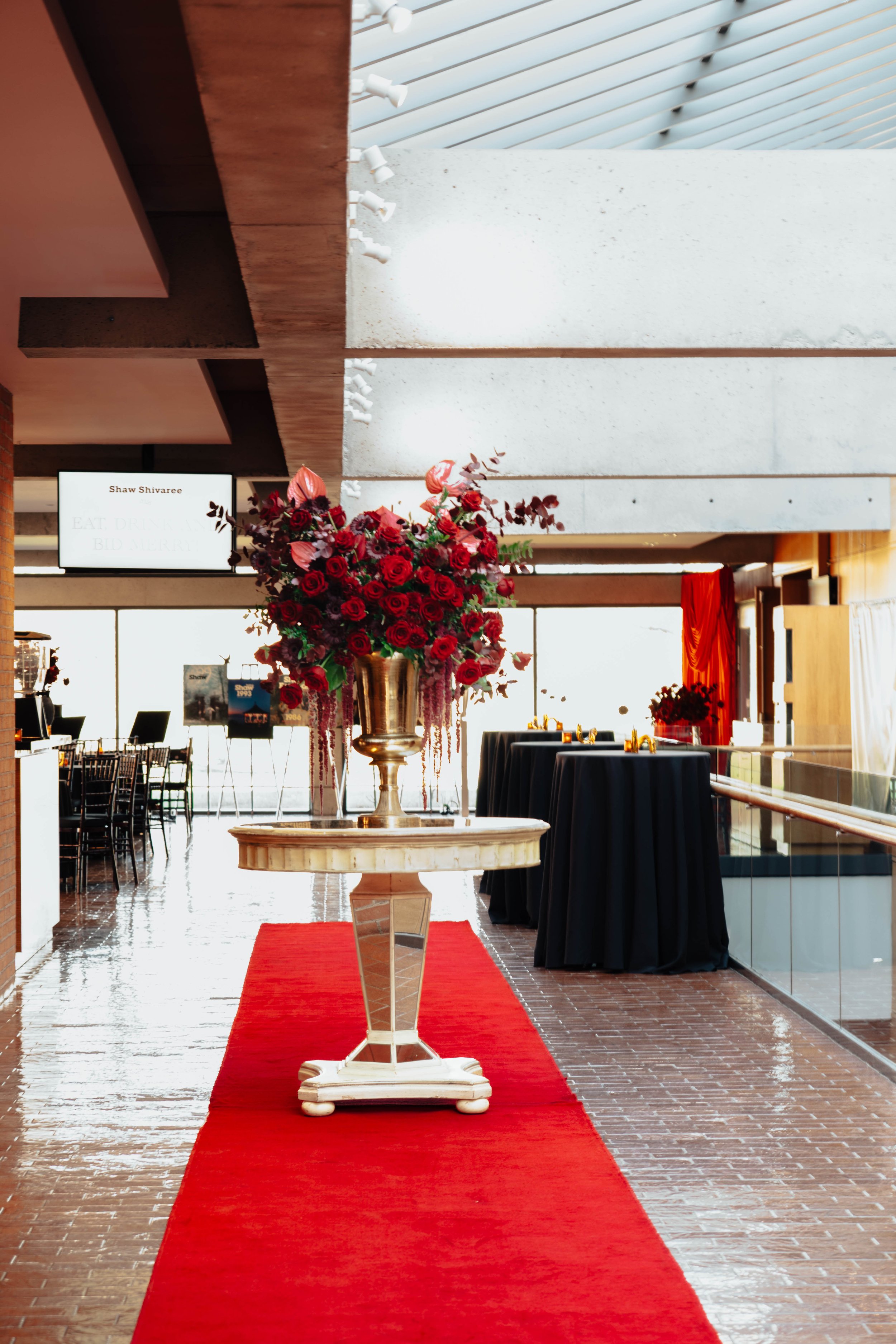 A large floral arrangement of red roses and pink flowers in a gold vase on a marble table with a red carpet underneath, inside a modern building with high ceilings and large windows.