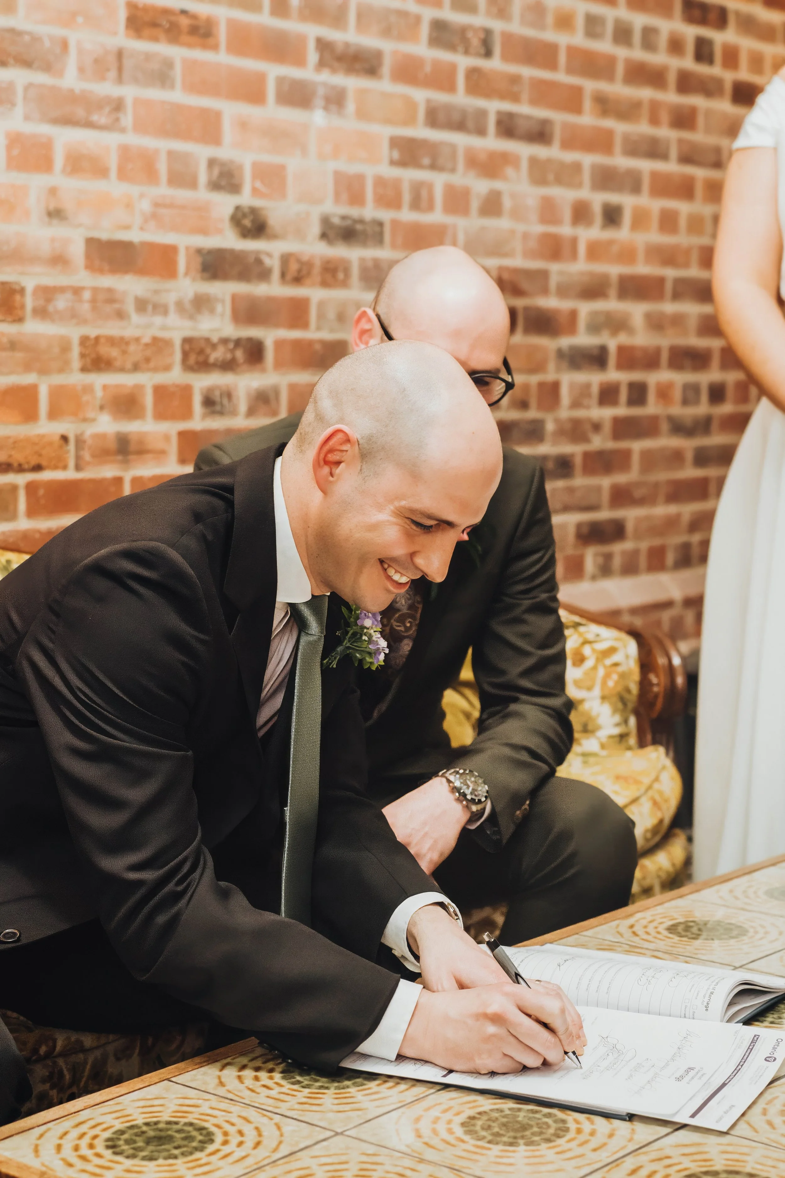 A man in a black suit and a tie signing a document at a table, with a smiling expression. An older man sits beside him, and a brick wall is in the background.