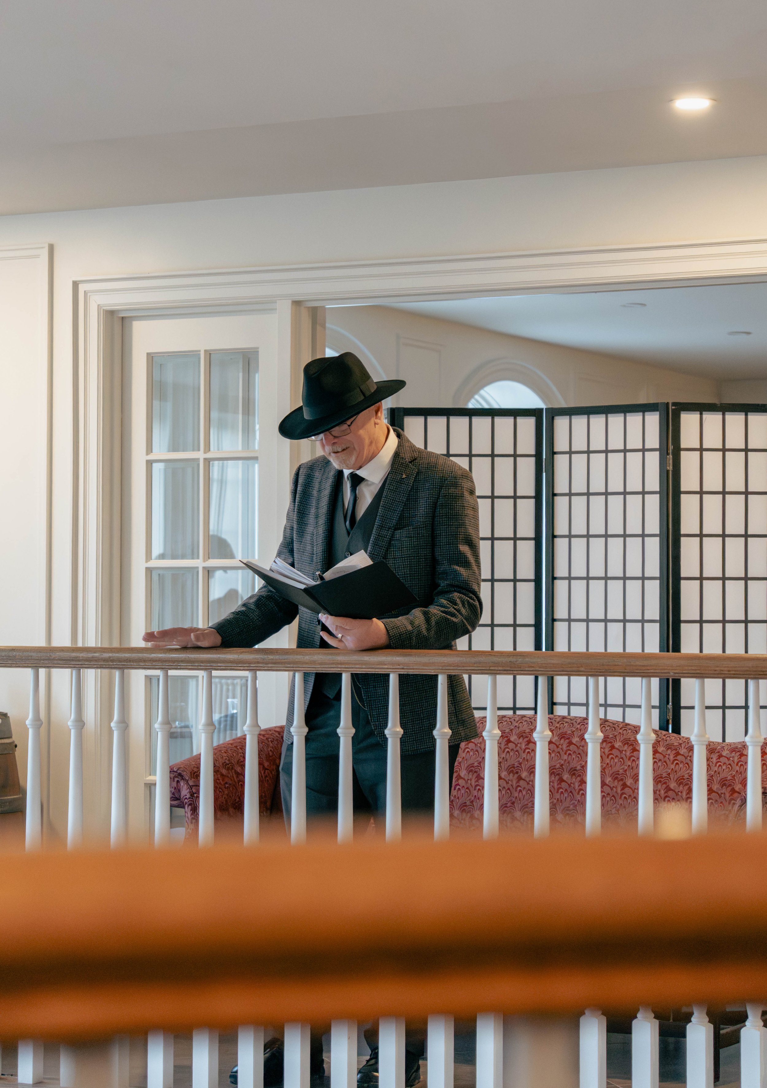 Older man wearing a hat, glasses, and a plaid suit jacket reading a book or script on a balcony.