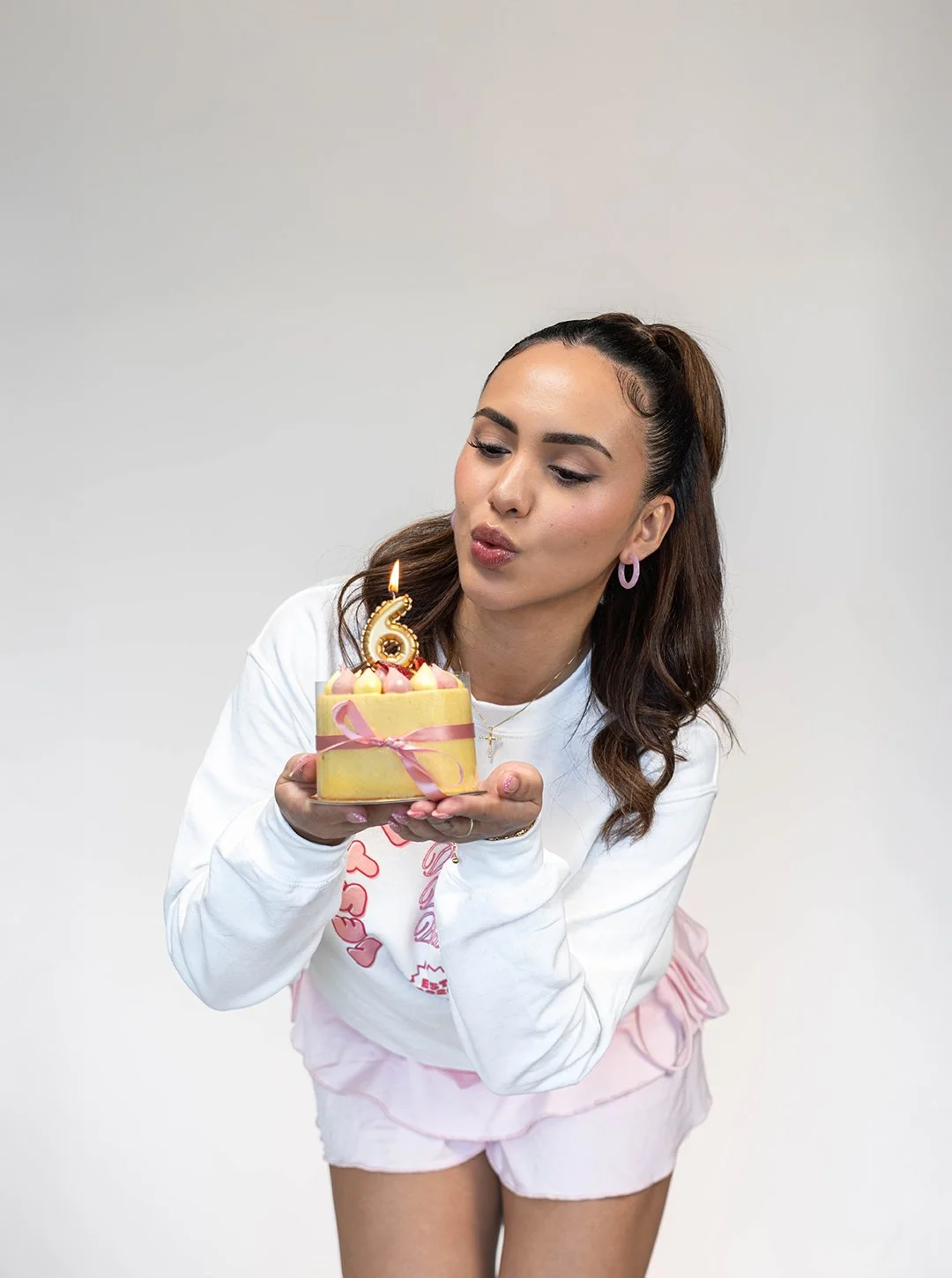 A woman with long brown hair, wearing a white long-sleeve shirt and pink shorts, is holding a small birthday cake with a lit candle shaped like the number 6. She is leaning forward, about to blow out the candle, with her lips puckered. The cake is de