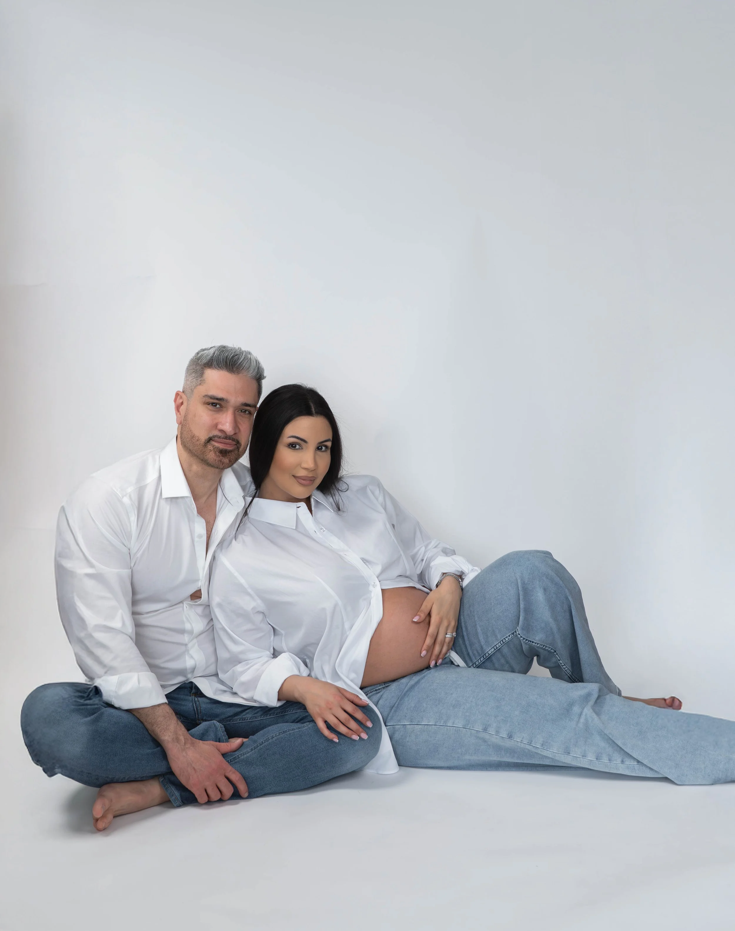 A man and a woman sitting together on the floor against a plain white background. The man has gray hair and a beard, wearing a white shirt and blue jeans. The woman has black hair, wearing a white shirt and blue jeans, with one leg bent and resting o