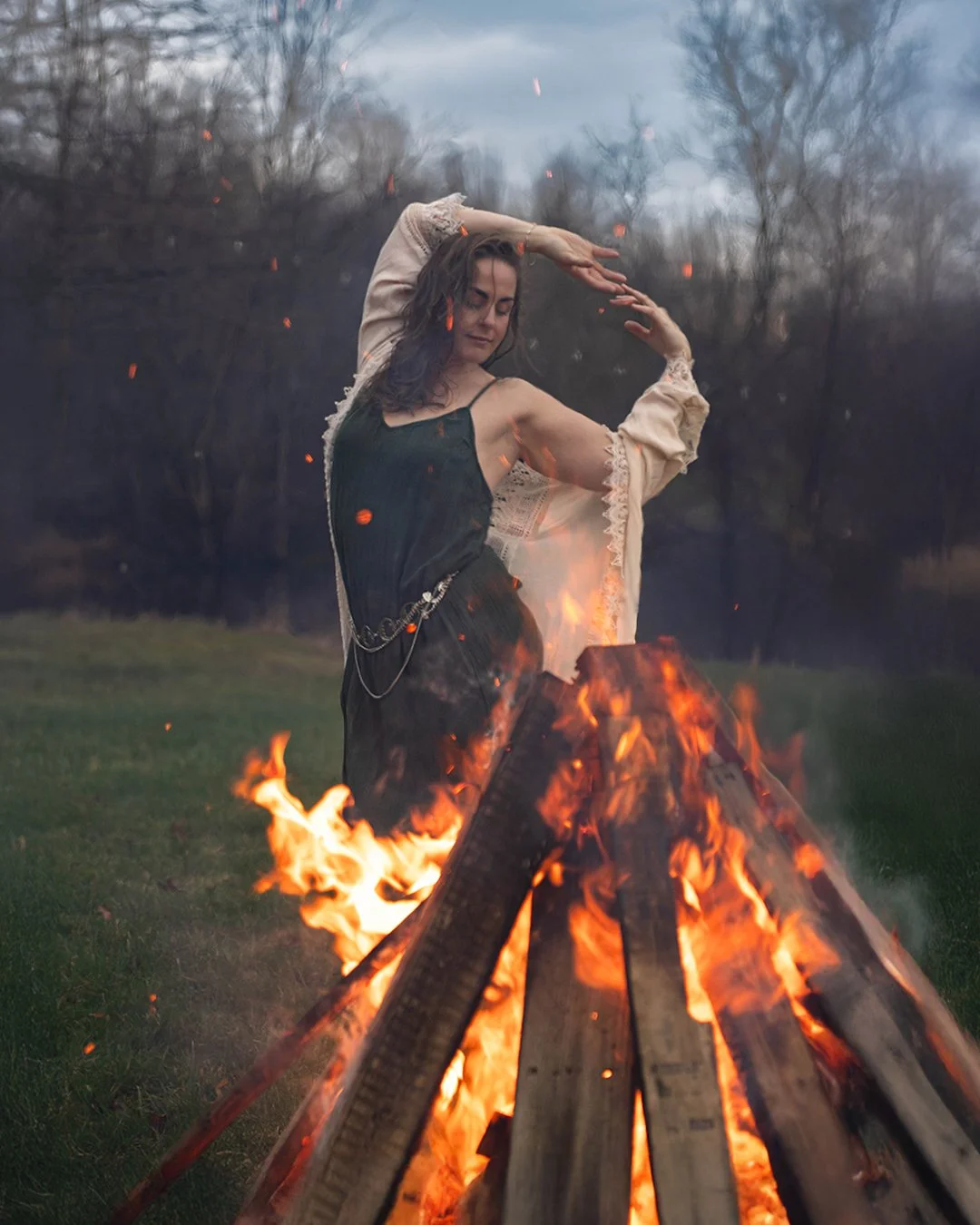 A woman standing near a large bonfire outdoors with her eyes closed and arms raised, wearing a black dress and a cream-colored shawl, trees and an overcast sky in the background.