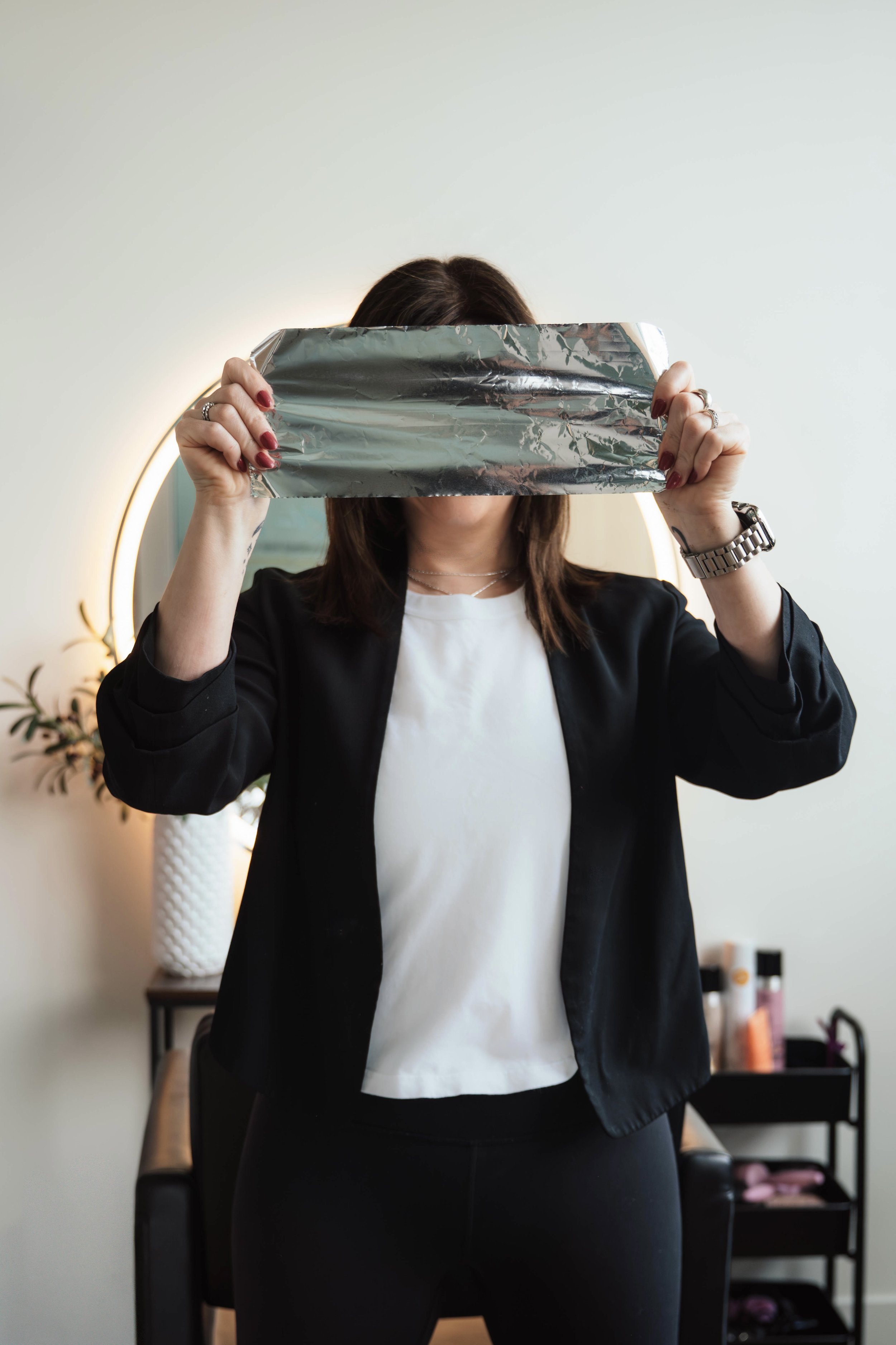 A woman with dark hair and wearing a black blazer and white shirt holds a silver foil package in front of her face, obscuring her identity. She is standing in a room with a mirror, a plant, and a cart with beauty products.