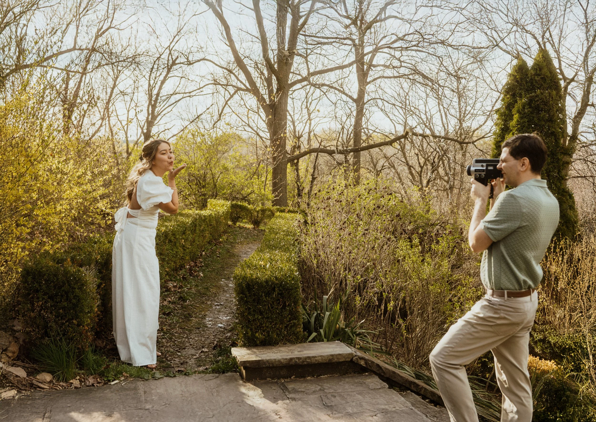 A woman in a white dress blowing a kiss while a man in beige pants and a grey shirt takes a photo of her with a camera in a garden with trees and bushes.