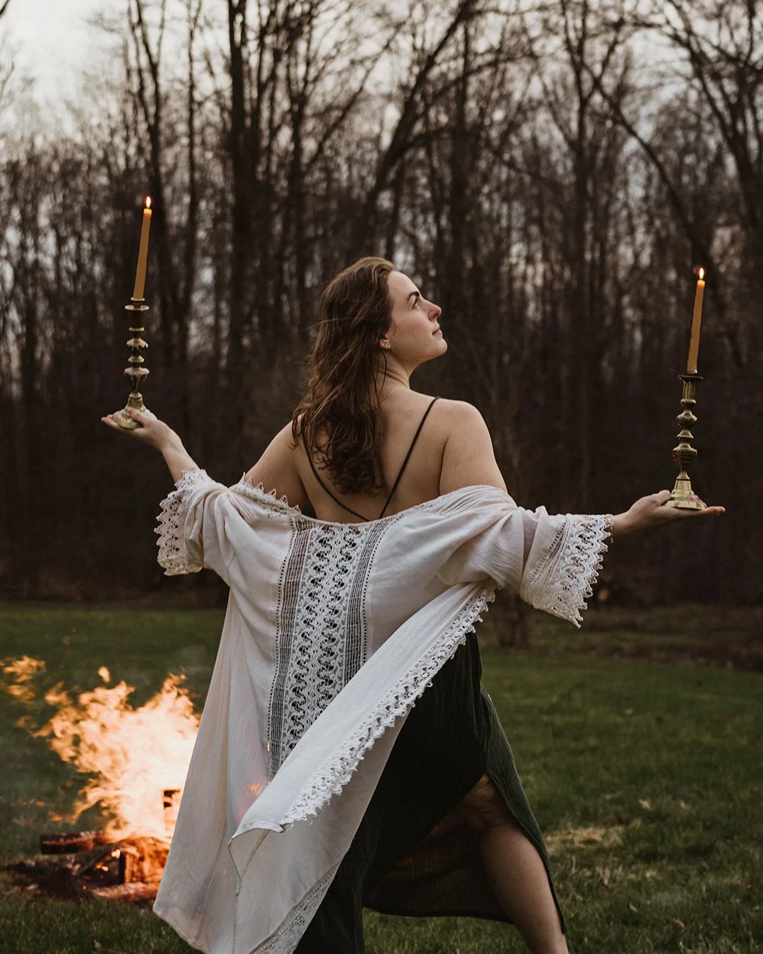 A woman standing in a grassy area at dusk, holding two lit candles on brass candlesticks, wearing a white embroidered shawl and a black dress with an open slit, with a small fire burning in the background and trees with bare branches behind her.