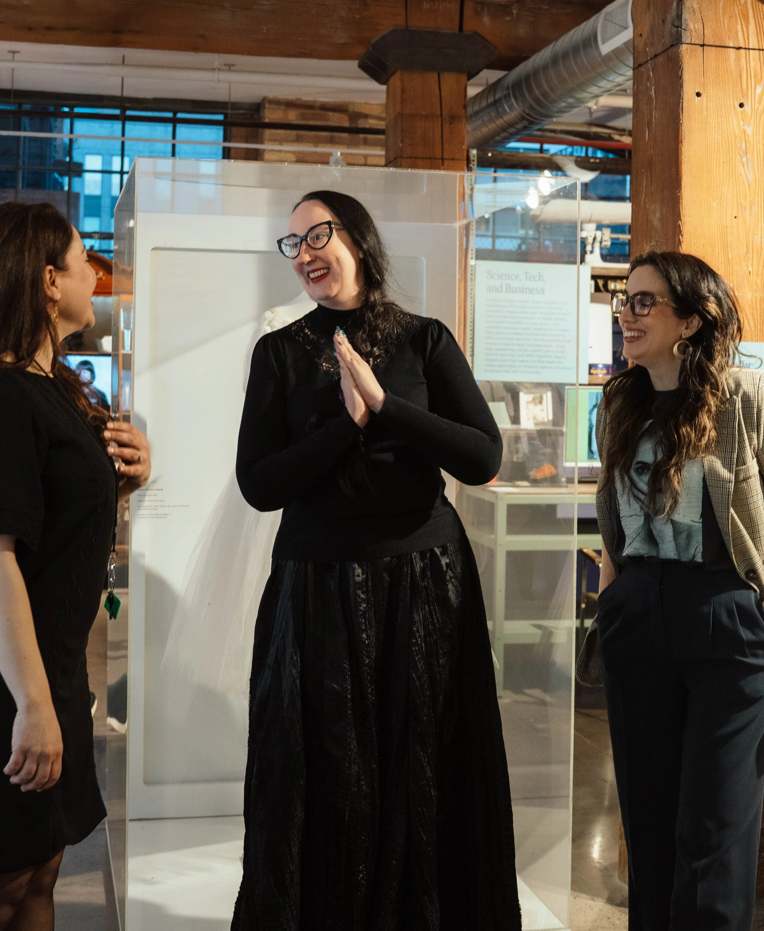 Three women smiling and talking in an indoor space with wooden beams and industrial elements. One woman is inside a transparent display case, wearing black, and the other two women are talking to her.