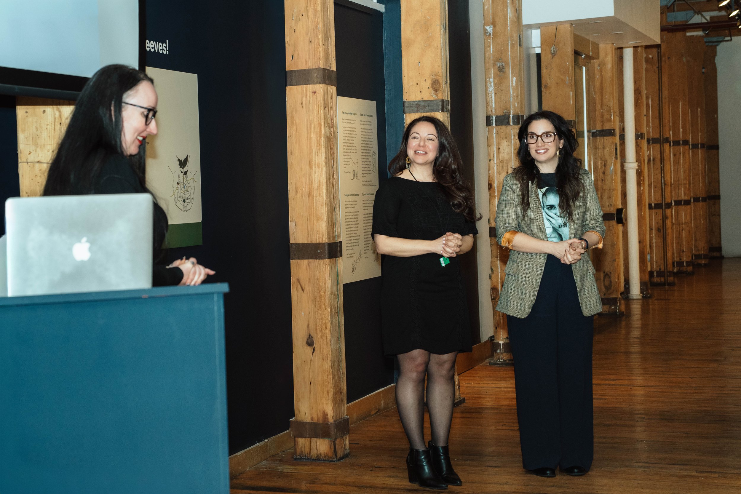 Three women standing and smiling during a presentation or event in a room with wooden walls and flooring.