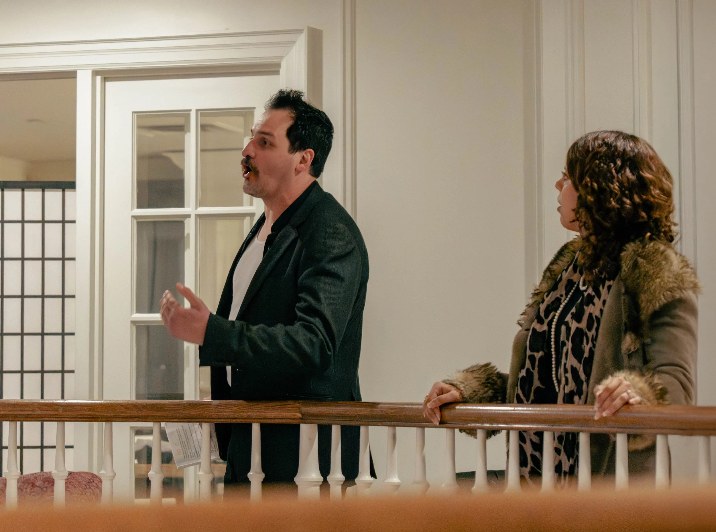 A man with dark hair and a mustache speaking to a woman with curly hair, both standing behind a wooden railing inside a house.