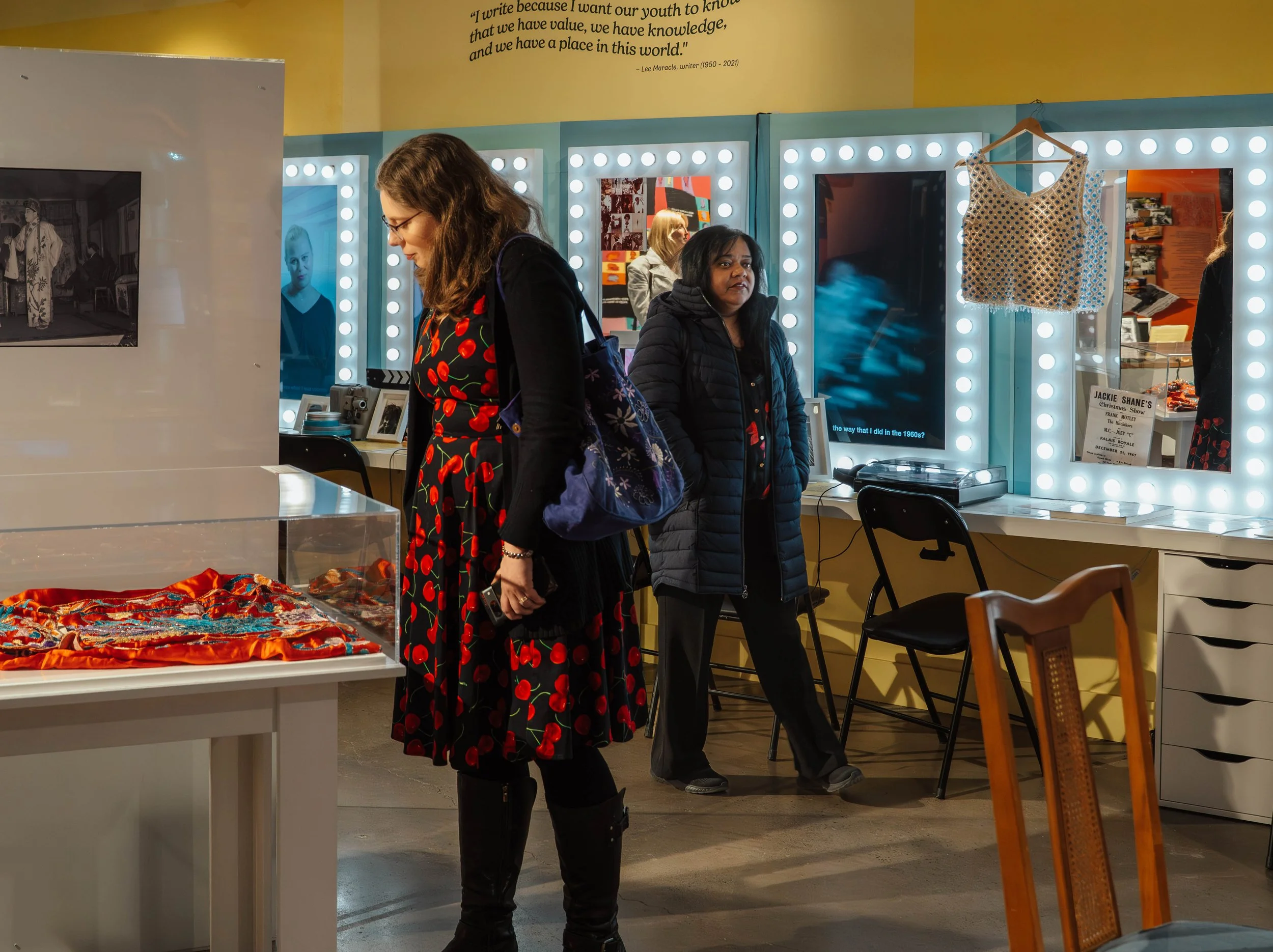 Two women in a museum exhibit, one in a cherry print dress looking at an object, and another in a dark jacket looking at the camera. There are illuminated mirrors, a hanging sleeveless top, and various items on display.