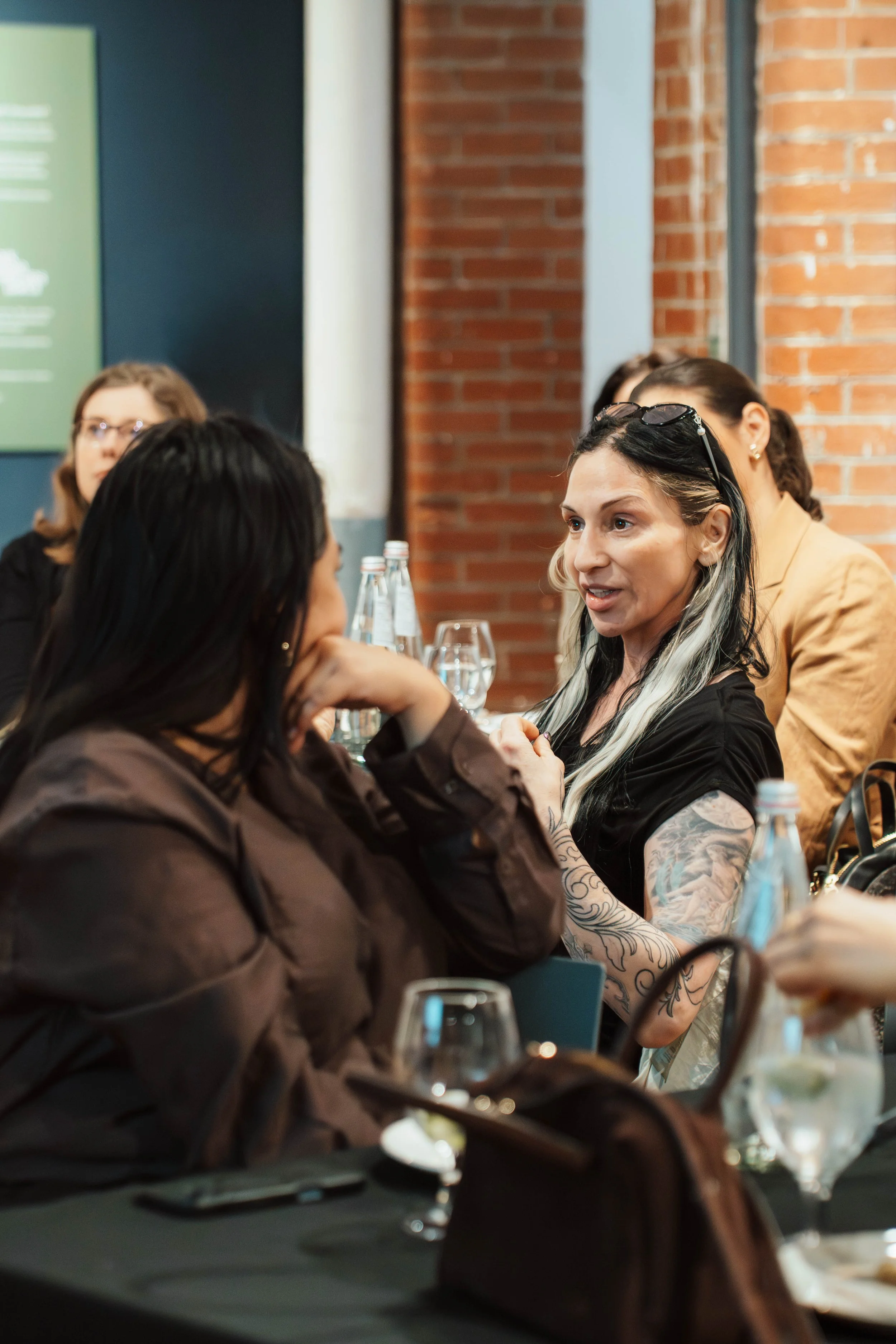 Women engaged in conversation at a table in a restaurant or conference setting with brick wall background.