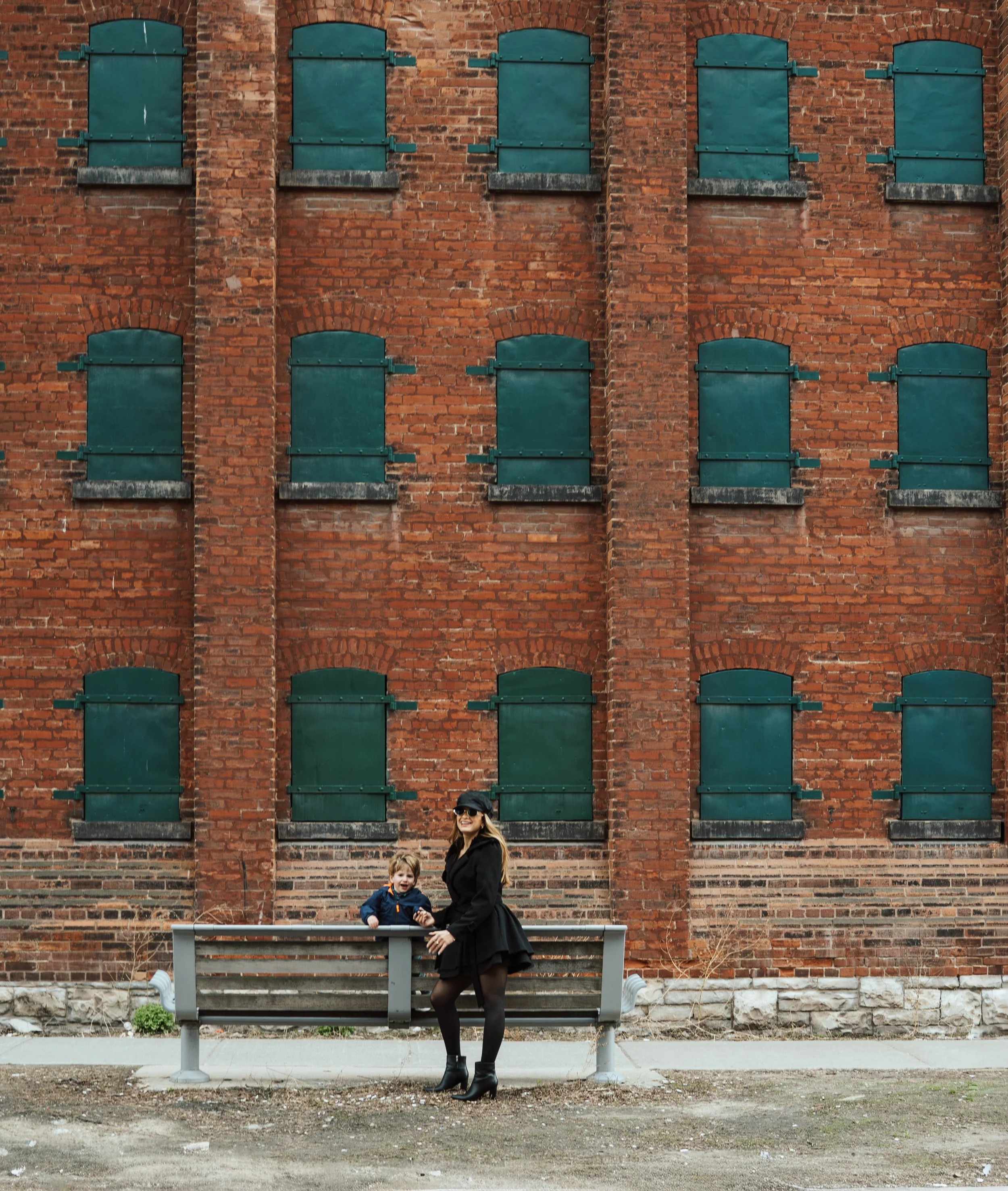 Woman and two children standing on a bench in front of a red brick building with boarded-up windows.