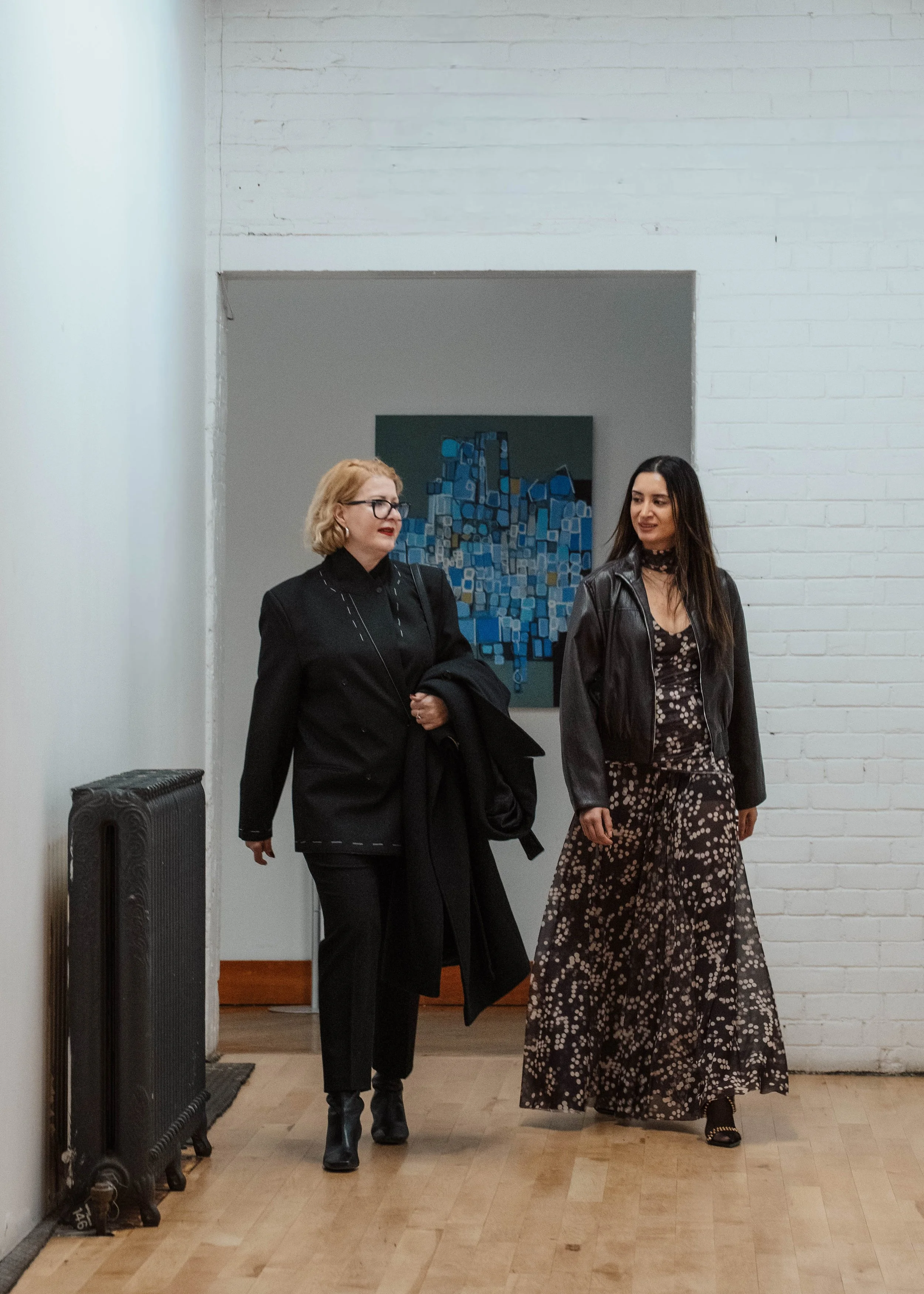 Two women walking and talking in an art gallery with white brick walls and a modern abstract painting in the background.
