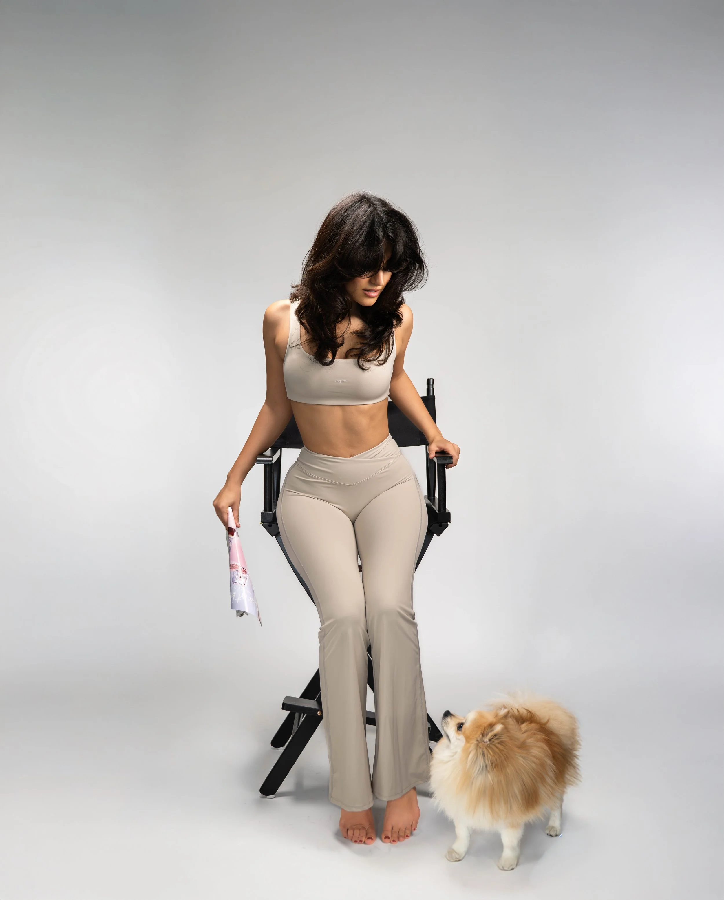 A woman in beige workout attire sitting on a director's chair, holding a magazine, with a small fluffy dog looking up at her against a plain white background.