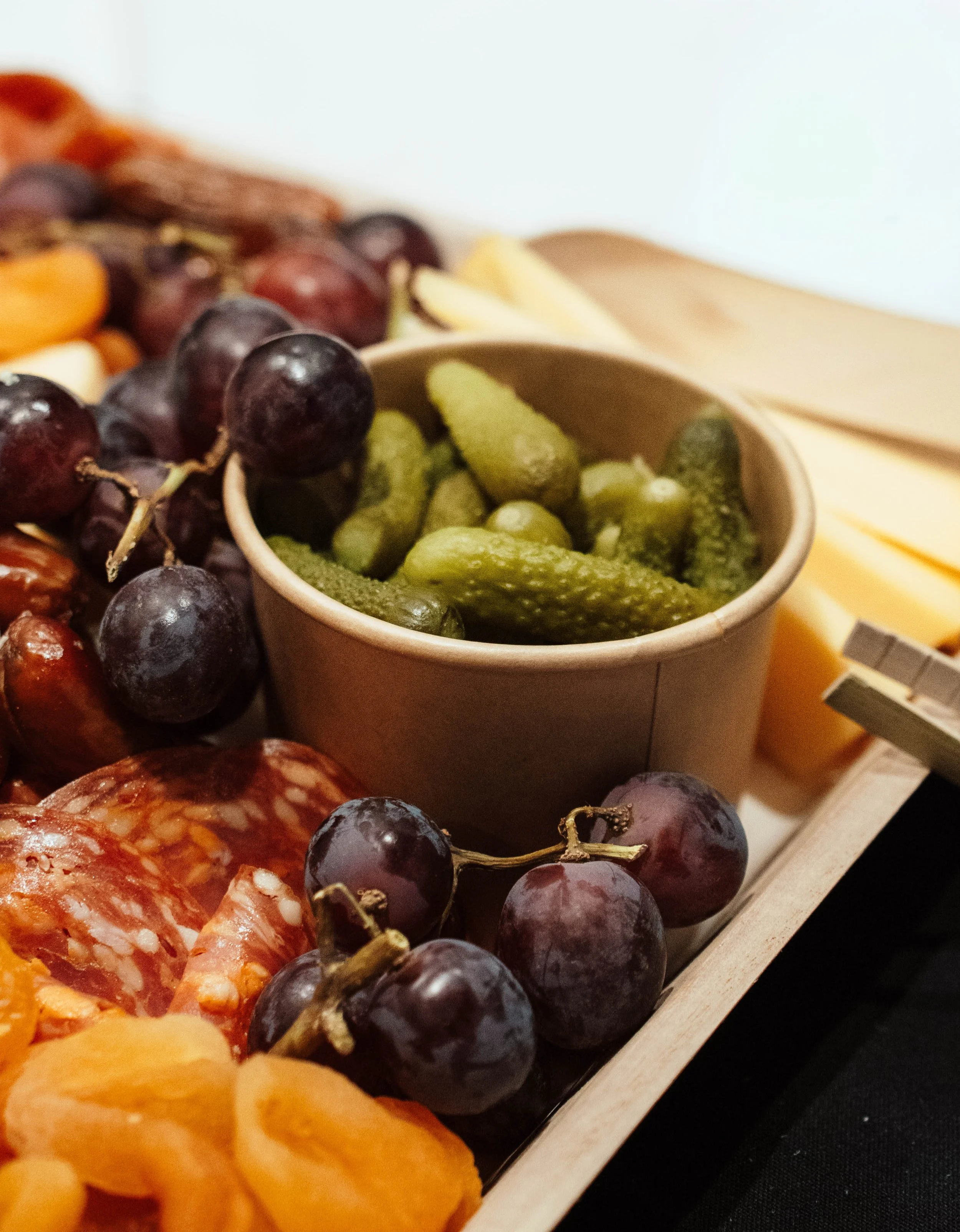 Close-up of a wooden serving tray containing grapes, pickles, sliced meats, cheese, and apricots.