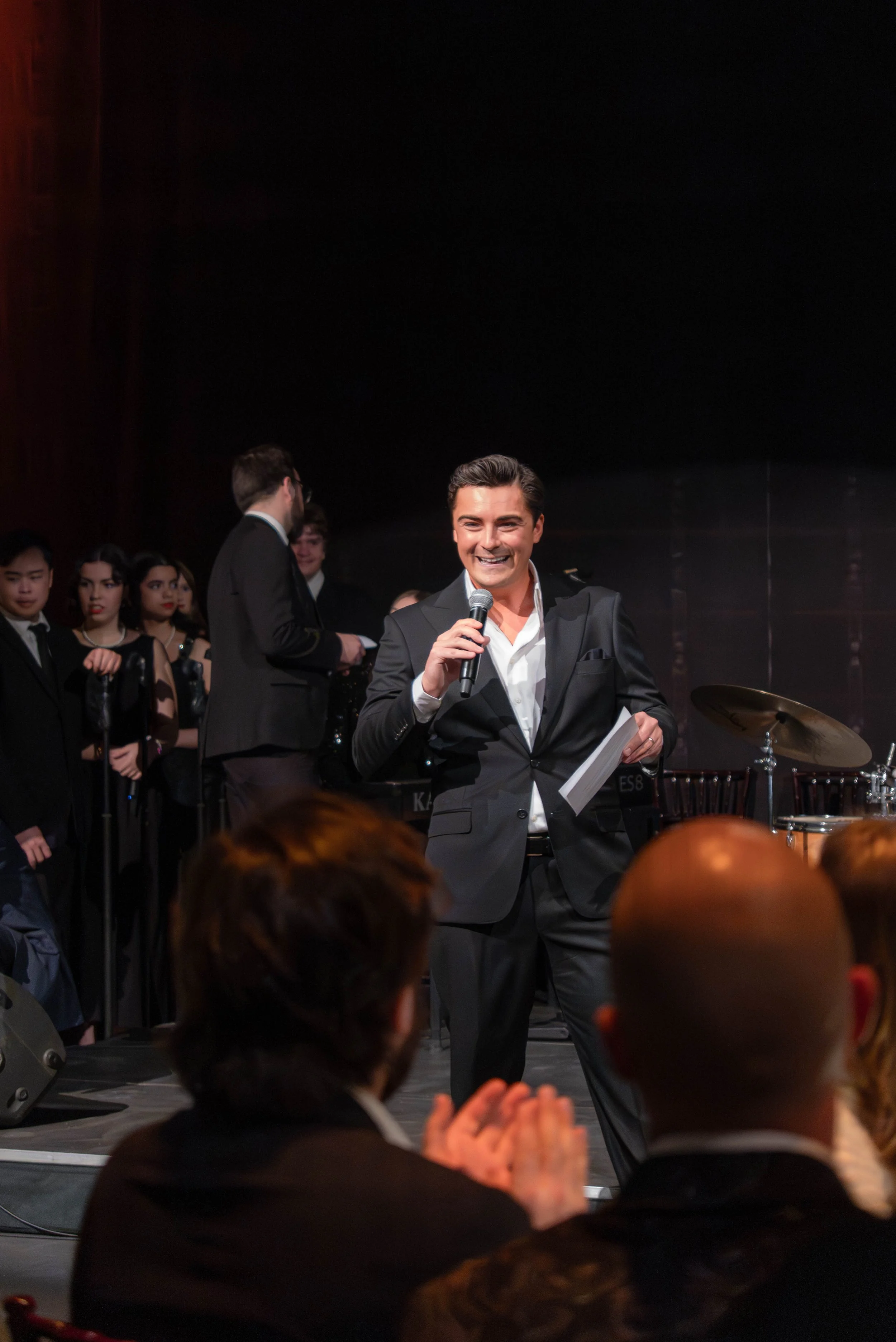 A man in a black suit smiling and speaking into a microphone at an event with a musical band and audience members in the background.