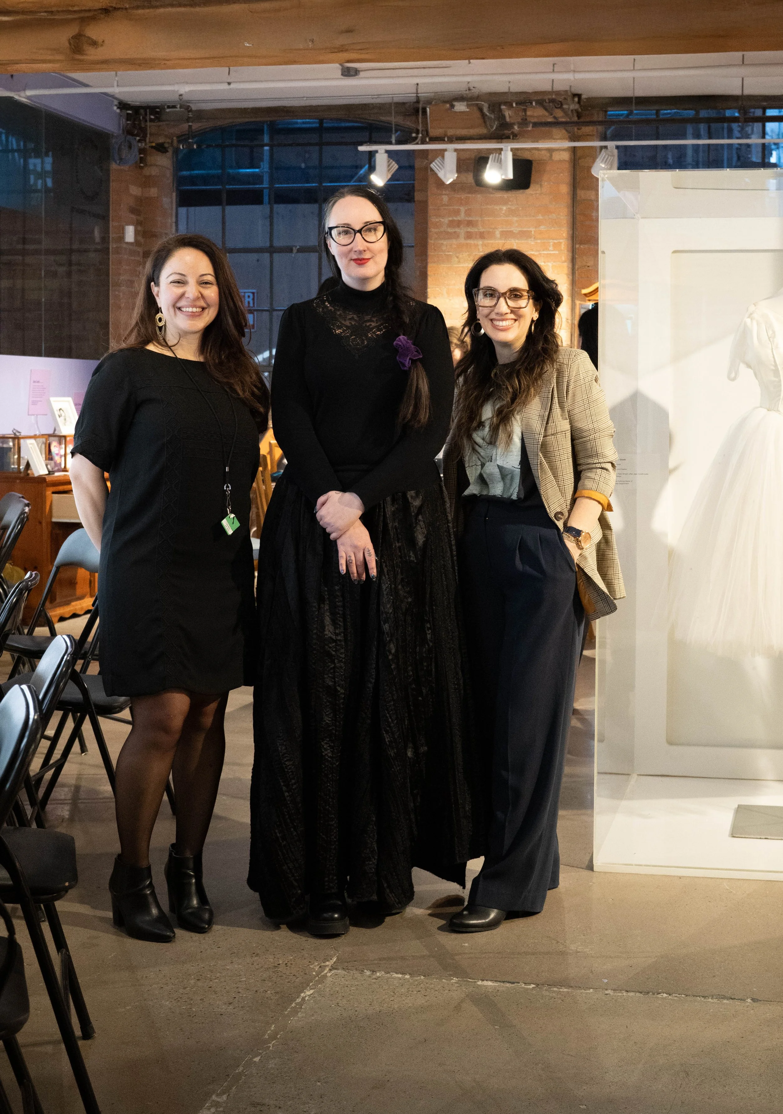 Three women standing together in an indoor setting with brick walls and a display case with a white dress behind them.
