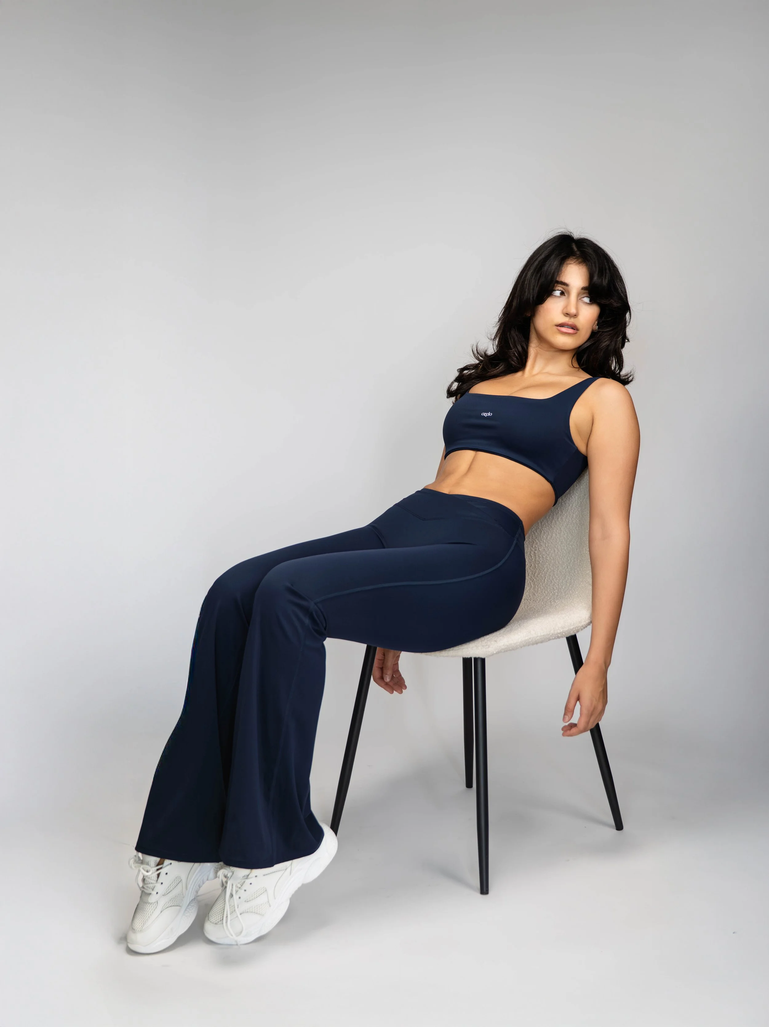 Woman sitting on a chair, wearing navy blue athletic wear and white sneakers, posing against a plain gray backdrop.