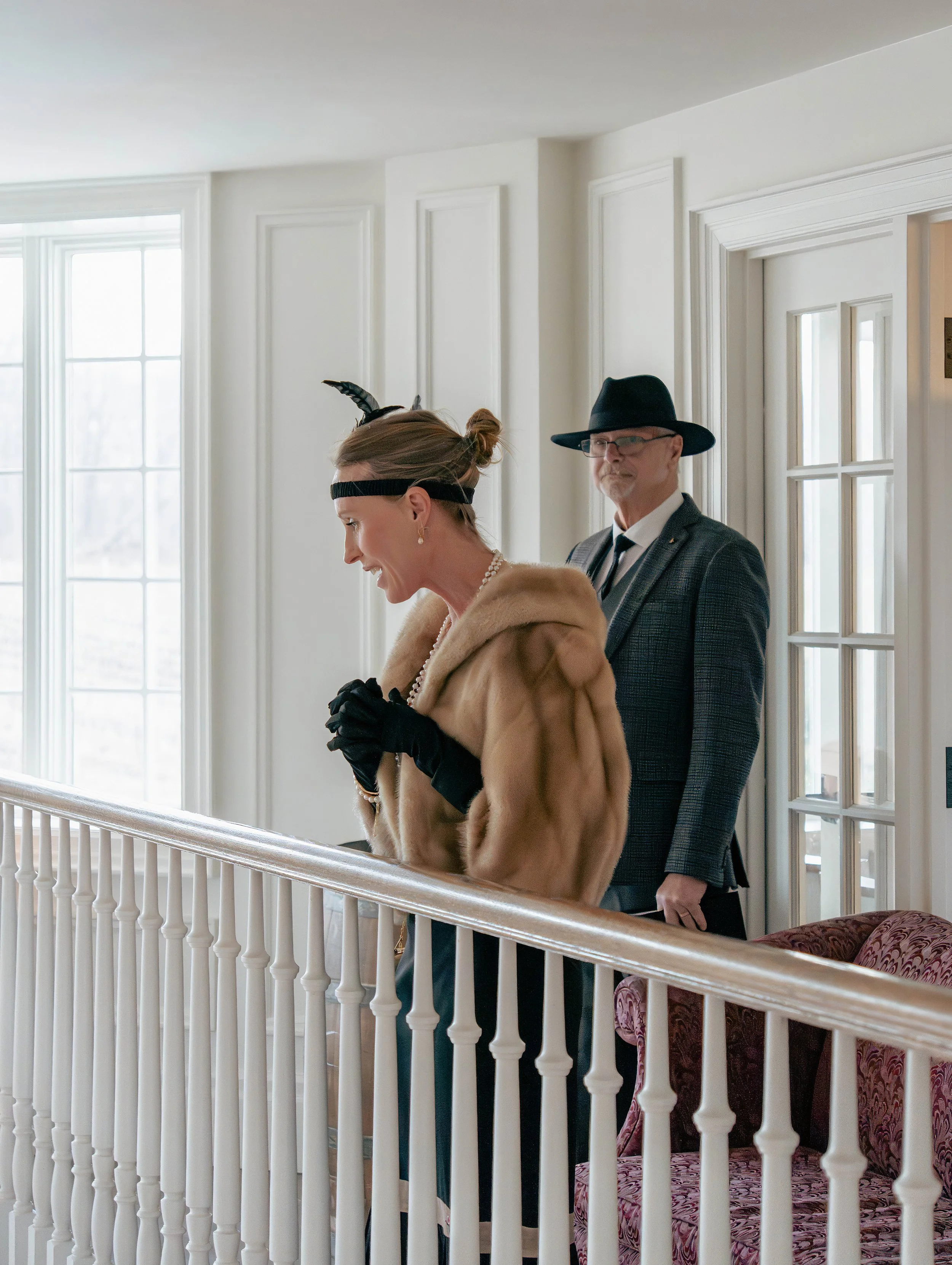 Woman in vintage clothing and accessories, including a fur coat, gloves, pearls, and a small hat, standing on staircase with man in suit and hat behind her.