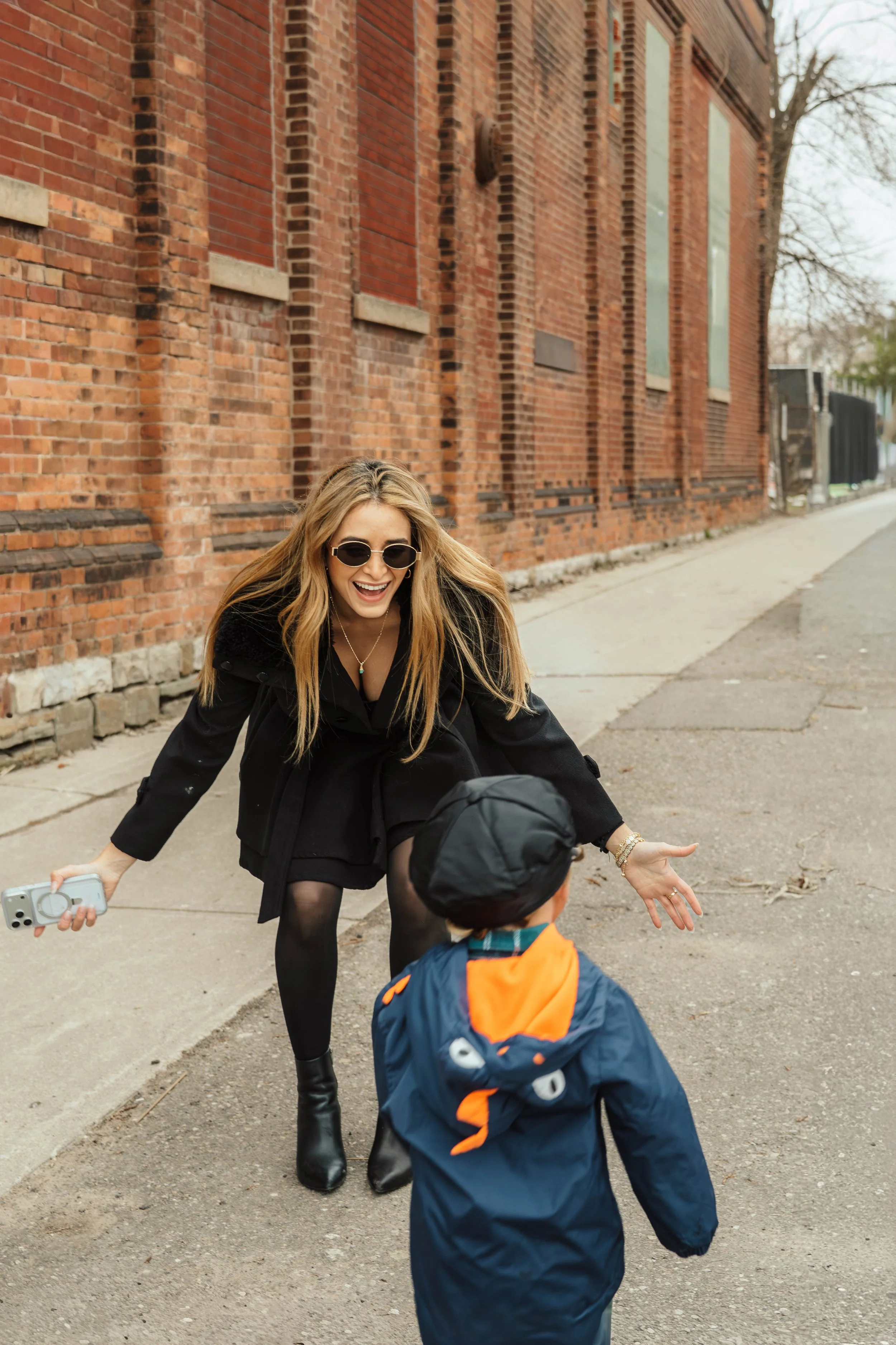 A woman with long blonde hair, wearing sunglasses, a black outfit, and black boots, is smiling and reaching out to a young boy wearing a blue and orange jacket, while standing on a sidewalk next to a brick building.