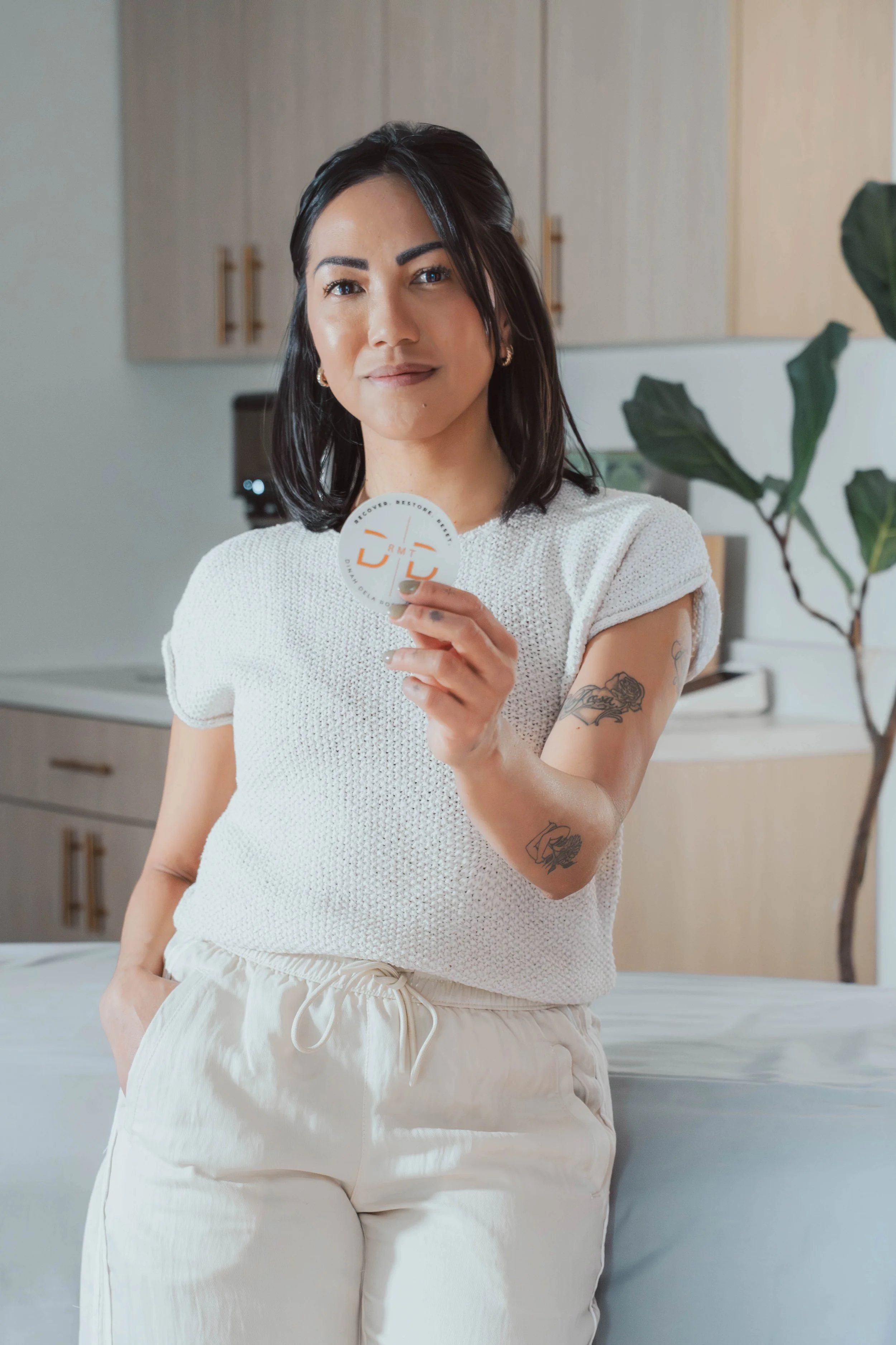 A woman with dark hair and tattoos on her arm, wearing a white sleeveless knit top and beige pants, standing in a room with wooden cabinets, holding a round sticker or token, smiling slightly.