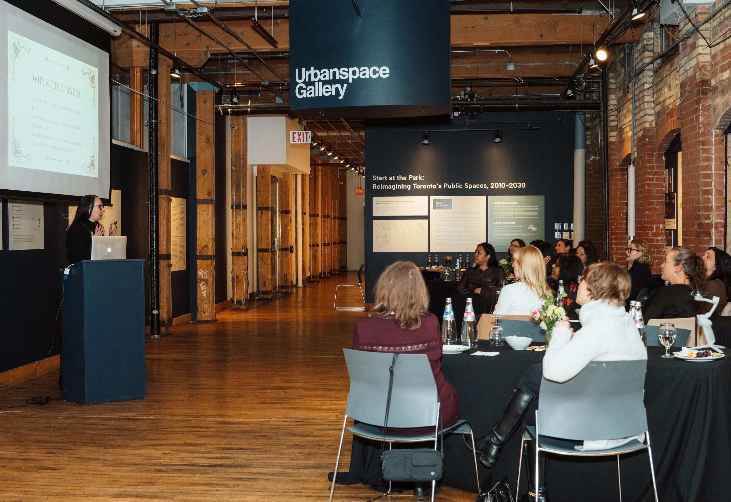 A woman giving a presentation at the Urbanspace Gallery while an audience attentively listens, seated at tables with bottles of water and plates of food, in a room with exposed brick walls and wood flooring.
