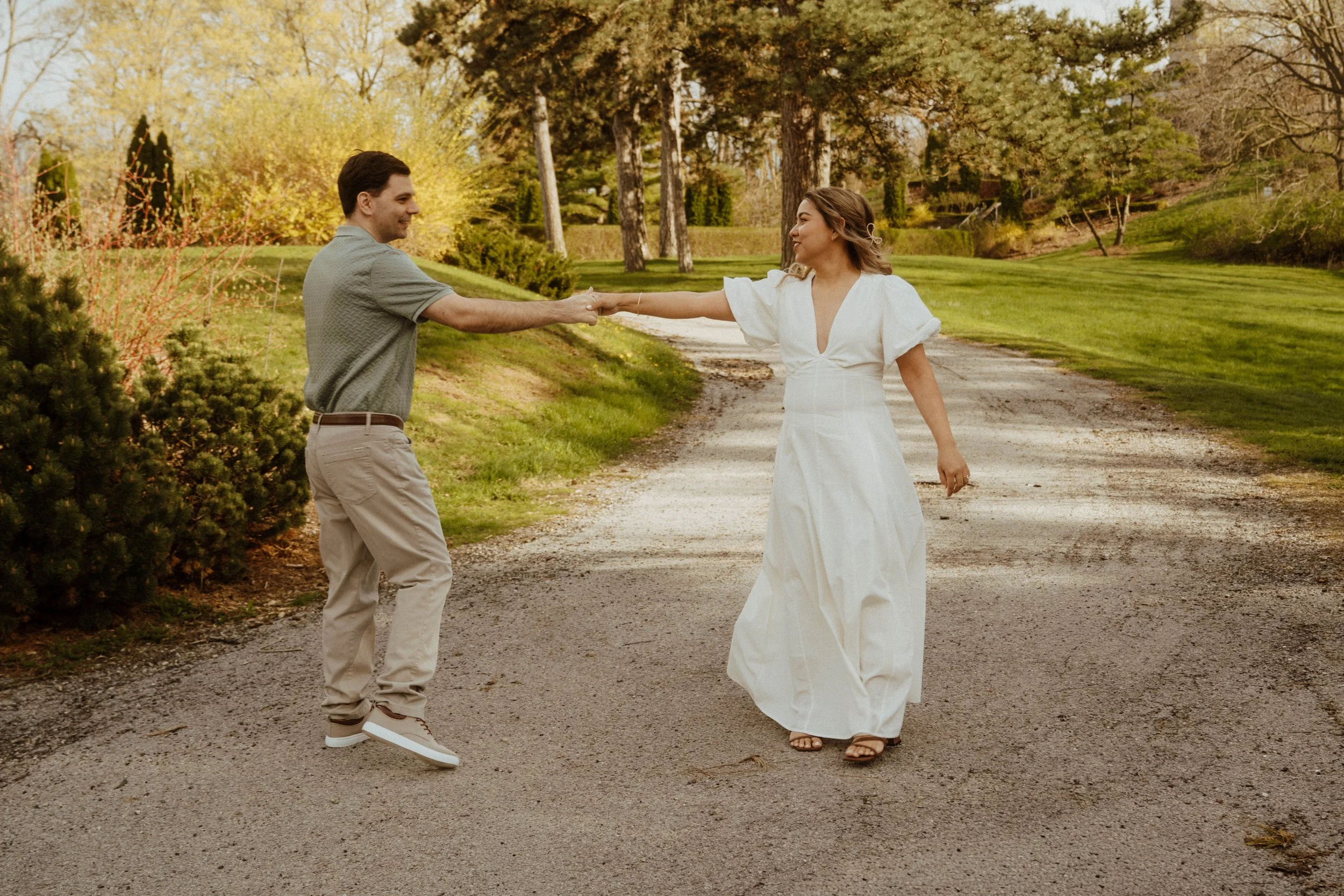 A man and woman are dancing outdoors on a dirt path surrounded by trees and greenery in a park or garden in the evening or late afternoon.